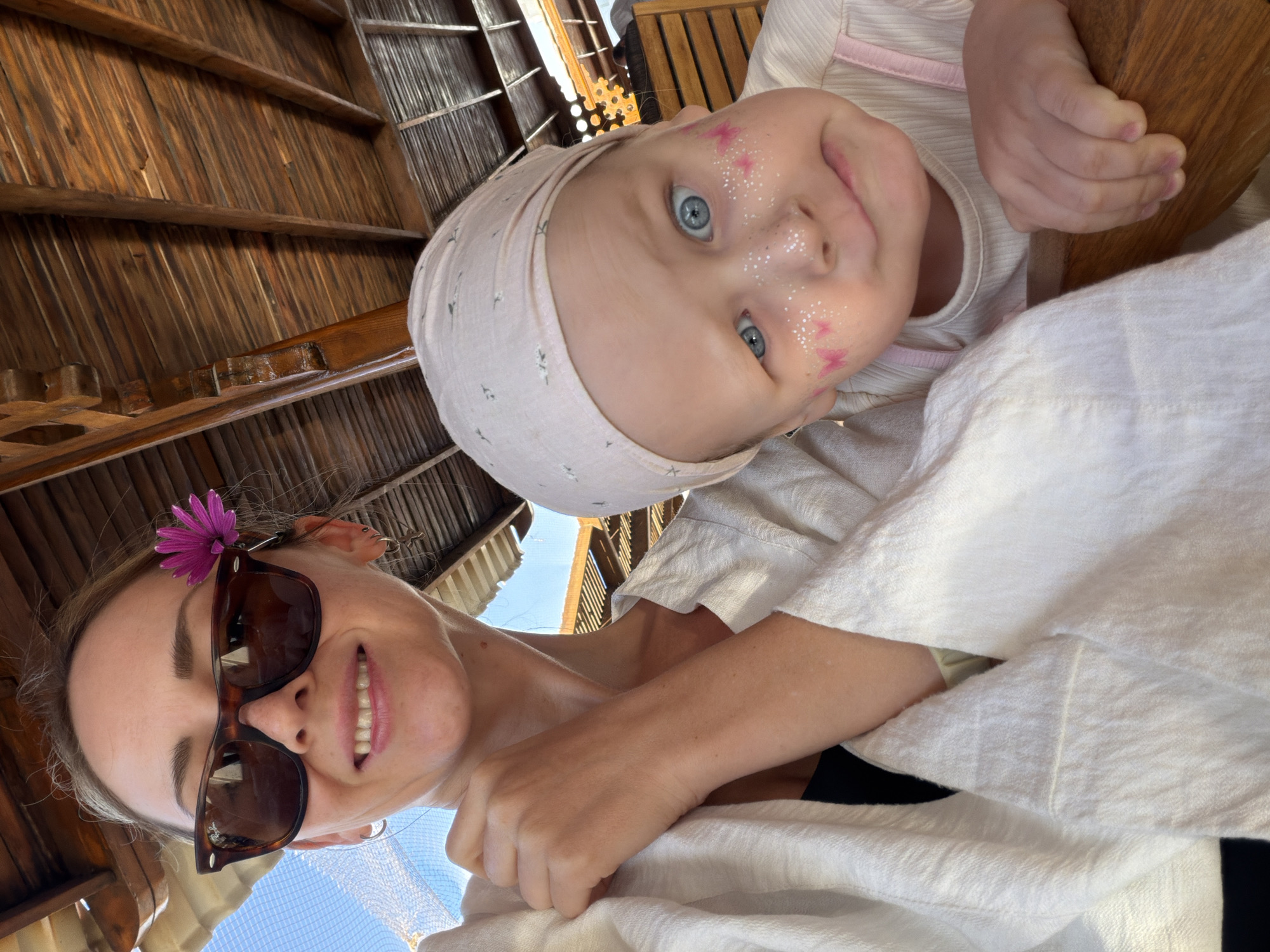 This media file captures a close-up, low-angle selfie of an adult woman and a baby, likely taken in a shaded, outdoor setting in Sharm El Sheikh, Egypt.
The primary subjects are a woman and a baby, positioned closely together and looking towards the camera. The woman is on the bottom right, smiling broadly with teeth showing, wearing dark brown sunglasses. A vibrant purple flower is tucked behind her left ear. She wears a light-colored, possibly white, garment, and her arm is visible.
Above and slightly to the left of the woman is the baby, with striking blue eyes and a light pink or beige head covering adorned with subtle patterns. The baby's face is decorated with white glitter and small pink butterfly-like shapes on the cheeks and nose, giving a playful appearance. The baby's small hand is visible, holding onto a wooden object. The baby has a curious or slightly surprised expression, with a hint of a smile.
The setting appears to be under a wooden structure with a thatched or slatted roof, possibly a cabana, pergola, or shaded area, consistent with a warm climate like Sharm El Sheikh. Sunlight peeks through parts of the roof and the blue sky is visible in the background, indicating daytime and clear weather. The overall mood is joyful and relaxed. No visible text is present in the image.