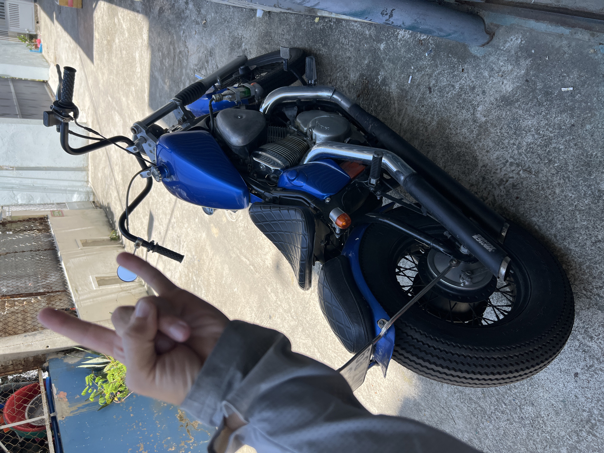 The image depicts a blue custom motorcycle, styled as a bobber or chopper, parked on a concrete alleyway in what is identified as Nha Trang, Vietnam. In the immediate foreground, a person's left hand, wearing a gray long-sleeved shirt cuff, makes a "rock on" hand gesture, obscuring the upper left portion of the motorcycle's handlebars.
The motorcycle features a metallic blue fuel tank and rear fender. It is equipped with a black, diamond-stitched solo seat and a matching smaller passenger pad. The handlebars are black, ape-hanger style, with a visible brake fluid reservoir on the right side and a small, round chrome mirror on the left. The engine components are a mix of chrome and silver-colored metal, with "Honda" possibly visible on the crankcase cover. A small, clear bottle containing a green liquid is secured near the engine. The exhaust system features chrome headers transitioning to black mufflers. The rear wheel has black spokes and a thick, heavily-treaded tire.
The environment consists of a light gray concrete pavement, showing some wear and scattered cigarette butts. To the left of the motorcycle, a blue metal gate or door is partially visible, behind which is a chain-link fence and green foliage, including what appears to be an agave plant, and a red bucket. To the right and in the background, light-colored stucco building walls with a white metal gate are visible, indicative of residential structures. Strong sunlight illuminates the scene, casting distinct shadows. The motorcycle is stationary, and no other individuals are visible.