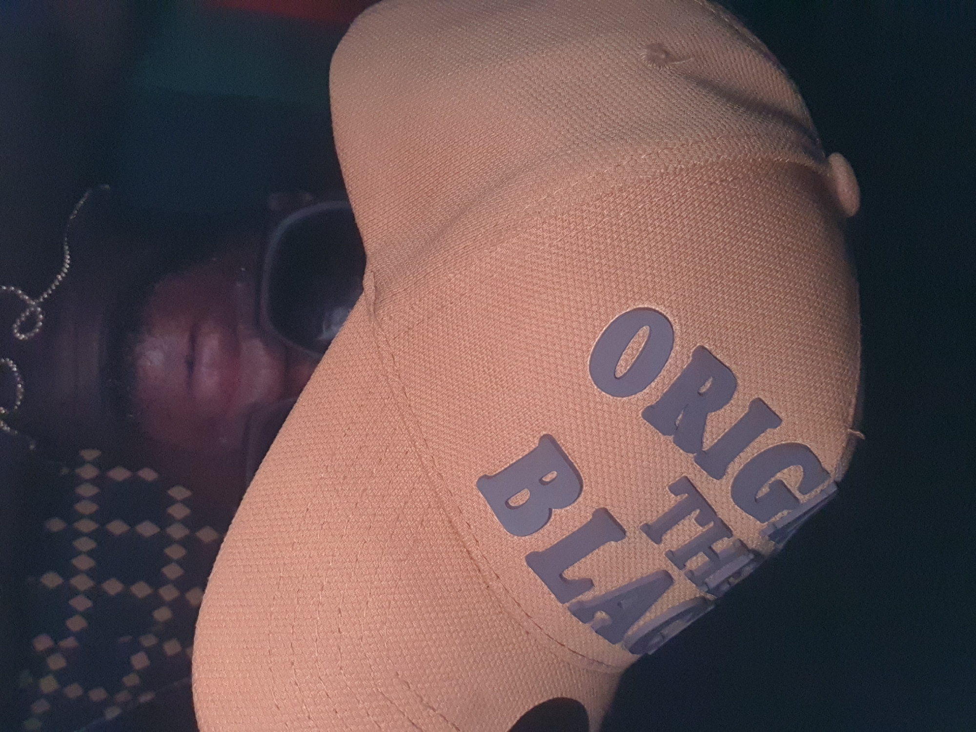 A close-up, slightly tilted, image focuses on a light orange baseball cap with the words "ORIGINAL THE BLACK" in gray lettering. A person is partially visible in the background, wearing sunglasses and possibly a patterned shirt or jacket. The environment is dark and undefined, with the background appearing to be out of focus. There is no indication of location, therefore the description of the city of Lokoja, Nigeria cannot be inferred.