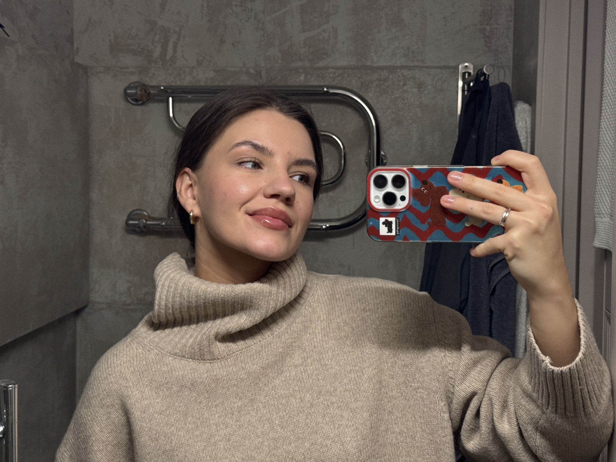 A young woman takes a mirror selfie in what appears to be a bathroom in Moscow, Russia. She has dark hair, which is pulled back, and her complexion is fair, with natural-looking makeup and glossy lips. She is wearing a light beige or tan ribbed turtleneck sweater and small gold hoop earrings. Her left hand, adorned with a silver ring on her ring finger and light pink nail polish, holds a smartphone up to capture the reflection. The phone's case is distinctive, featuring a red and blue zigzag pattern with cartoon bears or dogs. On the top edge of the phone case, the word "palogong" is visible, along with a small square design containing the number "1."
The background consists of a dark grey wall, possibly concrete or large tiles, giving the space a modern aesthetic. A chrome, curved heated towel rail is mounted on the wall behind her. To the right, a dark blue or black towel hangs on a hook, with another lighter grey towel partially visible. The lighting is somewhat dim, suggesting an indoor setting. The woman looks slightly to her right, with a soft, pleasant expression. There are no direct visual cues in the image itself to confirm the Moscow location beyond the provided context.