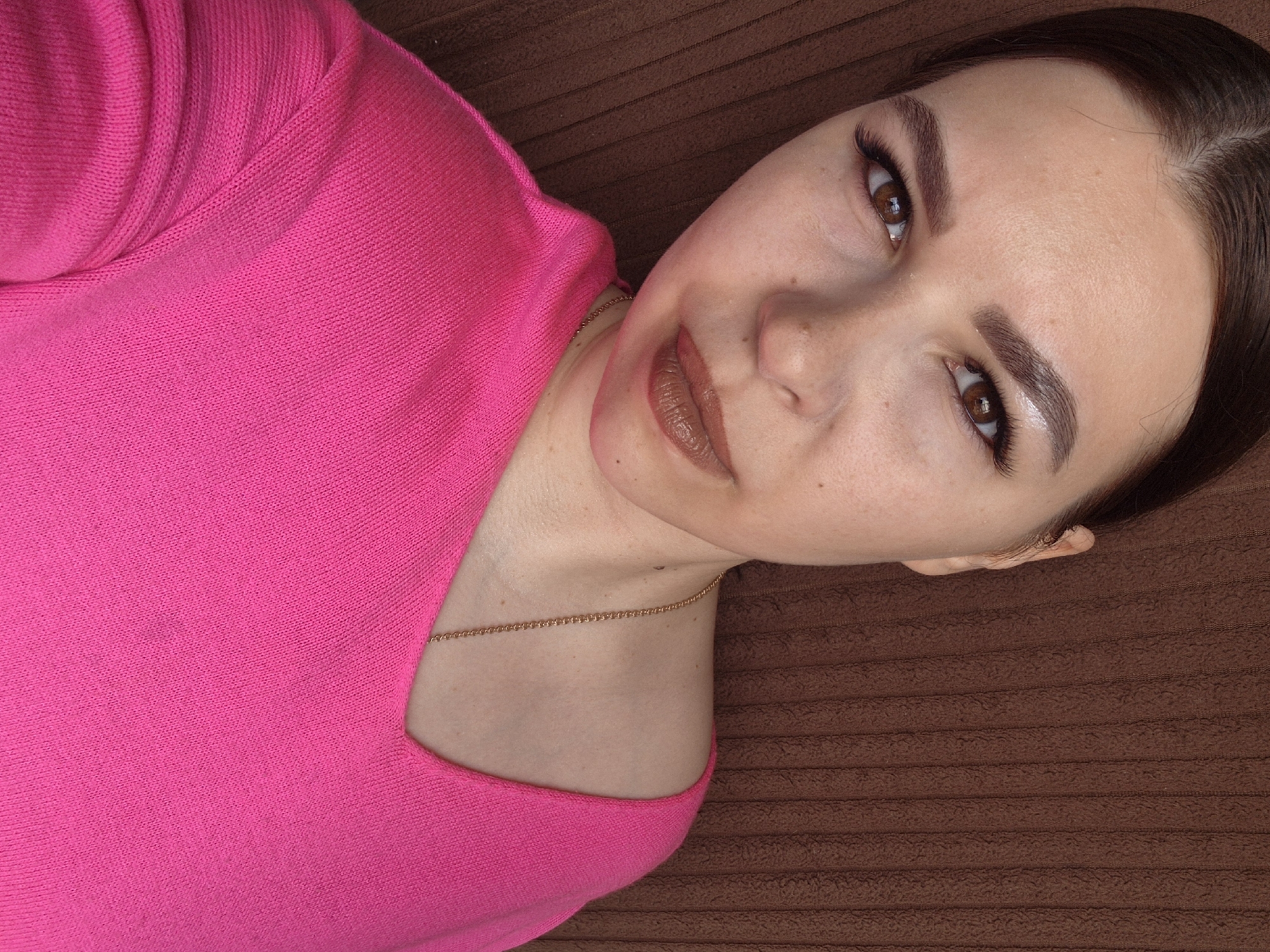 A medium, eye-level, close-up shot shows a light-skinned woman wearing a bright pink top and a delicate gold chain, lying on a dark brown, horizontally-striped surface. She has dark brown hair pulled back from her face, defined eyebrows, brown eyes, and glossy brown lipstick. The background is a close-up of a brown, horizontally-lined surface. There is no indication of this being in Tobolsk, Russia.