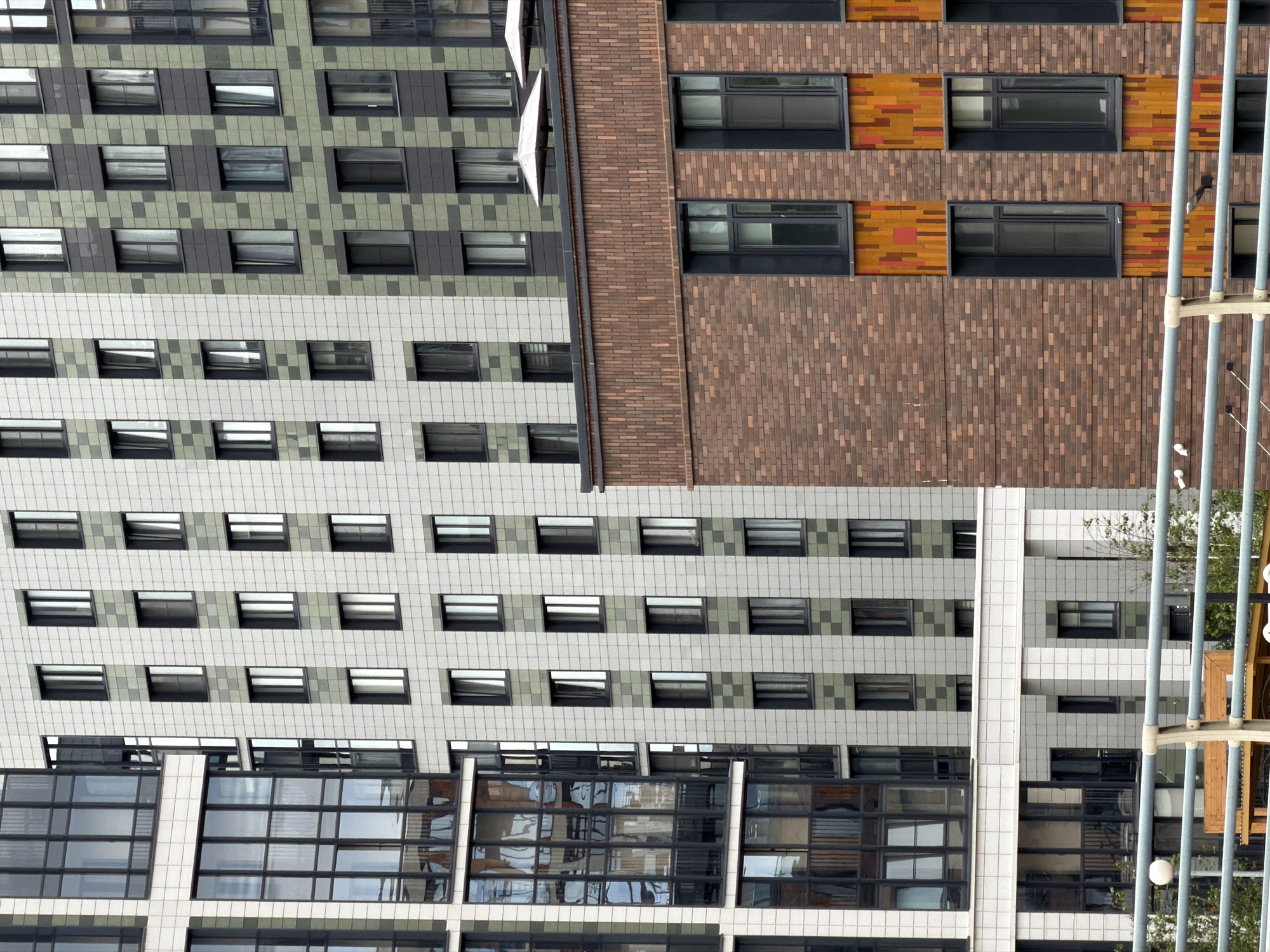 This image displays a section of a modern residential complex in Saint Petersburg, Russia, featuring several buildings with varied architectural styles. The leftmost building is characterized by a facade of large, dark-framed windows, suggesting a contemporary design with significant glass elements. Adjacent to it, a taller building exhibits a grid-like pattern of windows set against a light-colored tiled exterior, interspersed with vertical bands of green and grey tiles. Further to the right, another building showcases a similar tile pattern, but with a more prominent use of green tiles in a checkerboard arrangement.
The foreground of the image is dominated by a lower building constructed with brickwork, featuring dark-framed windows and decorative vertical panels in warm orange and yellow hues. Above this brick structure, two white umbrella-like structures are visible, possibly indicative of a rooftop amenity area. A metal railing runs across the bottom of the frame, obscuring some of the lower details of the buildings and suggesting a viewpoint from an elevated position, such as a balcony or another building. Vegetation and trees are subtly visible in the background, indicating an urban setting with some green spaces. The sky is overcast, contributing to a soft, diffused light that illuminates the scene. No people are clearly visible in the image, and the focus is entirely on the architectural details of the buildings.