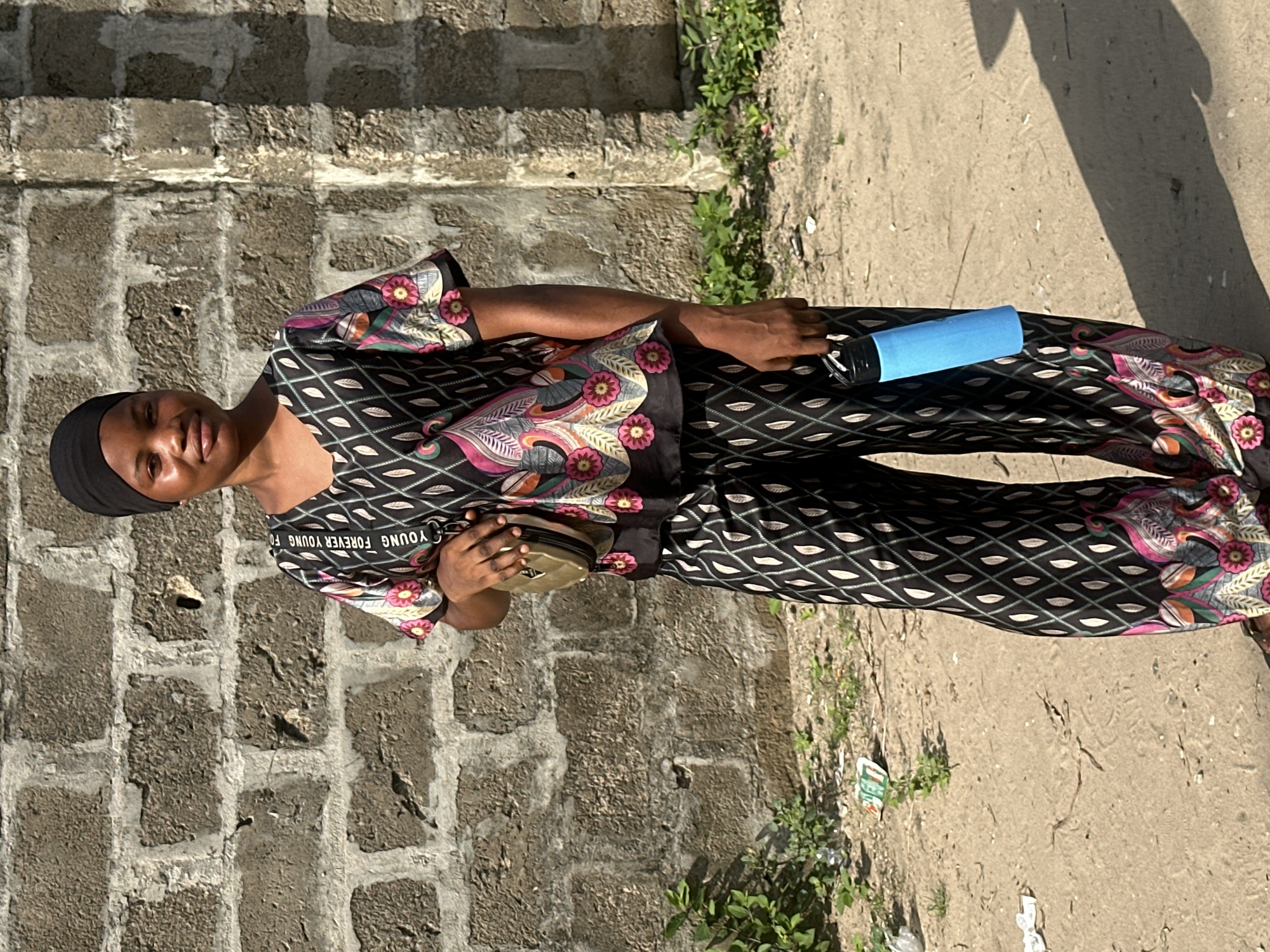 A young person stands outdoors in what appears to be Badagry, Nigeria, captured in bright daylight. The individual, likely female, is wearing a black head covering and a matching two-piece outfit. The attire features a black base with a repeating geometric pattern resembling white or light-colored leaves or diamonds, complemented by vibrant floral and paisley designs in shades of pink, green, and orange on the sleeves and the lower part of the pants. Text is visible on the chest area of the top, reading "YOUNG FOREVER YOUNG FO" (with the remainder likely "REVER").
In the left hand, the person holds a light blue insulated bottle or flask with a black cap. A small, light brown or tan pouch, possibly a fanny pack or cross-body bag, is held in the right hand. The individual is looking directly at the camera with a gentle smile, appearing calm and content.
The setting includes a rugged, multi-toned stone wall on the left side of the frame, possibly part of a building or compound. The ground is sandy or dirt-covered, with sparse green vegetation, likely weeds, growing along the base of the wall and in patches on the ground. Some small pieces of litter are visible on the ground. The presence of sharp, distinct shadows cast on the ground to the right indicates bright sunlight, suggesting it is likely mid-morning or afternoon with clear weather.