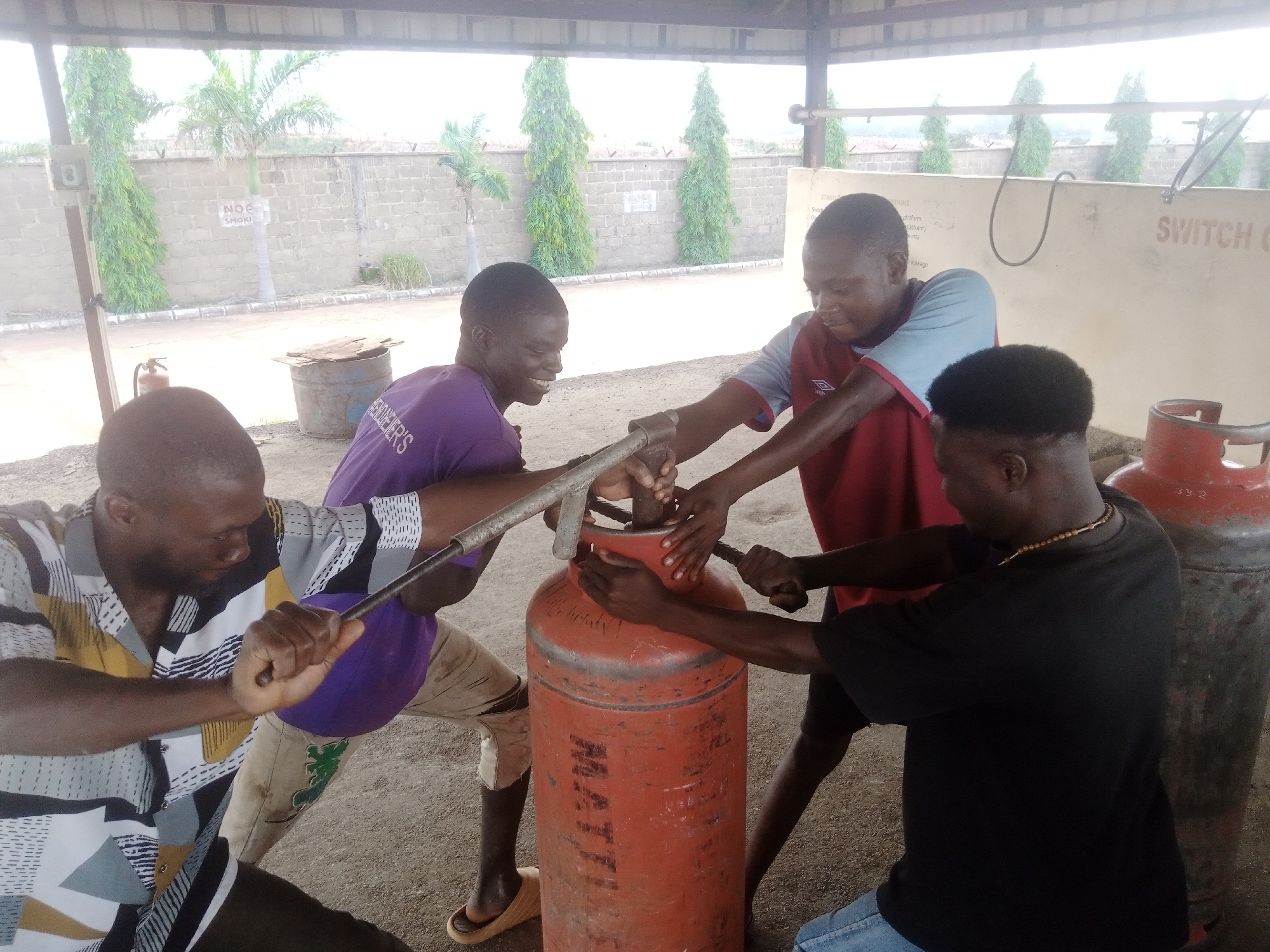Four young men are working together to move or position a large, red gas cylinder. They are gathered around the cylinder, with at least three of them gripping it or tools attached to it. One man in a patterned shirt is holding a metal bar that appears to be leveraged against the cylinder's valve. Another man in a purple shirt is also holding the metal bar. A third man in a red and blue shirt is holding onto the cylinder itself, while a fourth man in a black t-shirt is also touching the cylinder.
The setting appears to be outdoors under a covered structure, possibly a workshop or a storage area. The ground is dirt or gravel. In the background, there is a light-colored brick wall with some greenery, including palm trees and tall, slender trees. There is also a metal fence. On the wall, there is a sign that reads "NO SMOKING". Another wall in the background has text that includes "SWITCH ON".
The individuals appear focused on their task, and their expressions suggest exertion and concentration rather than distress. The lighting suggests it is daytime, likely a bright, possibly overcast day. The overall atmosphere is one of collaborative effort.