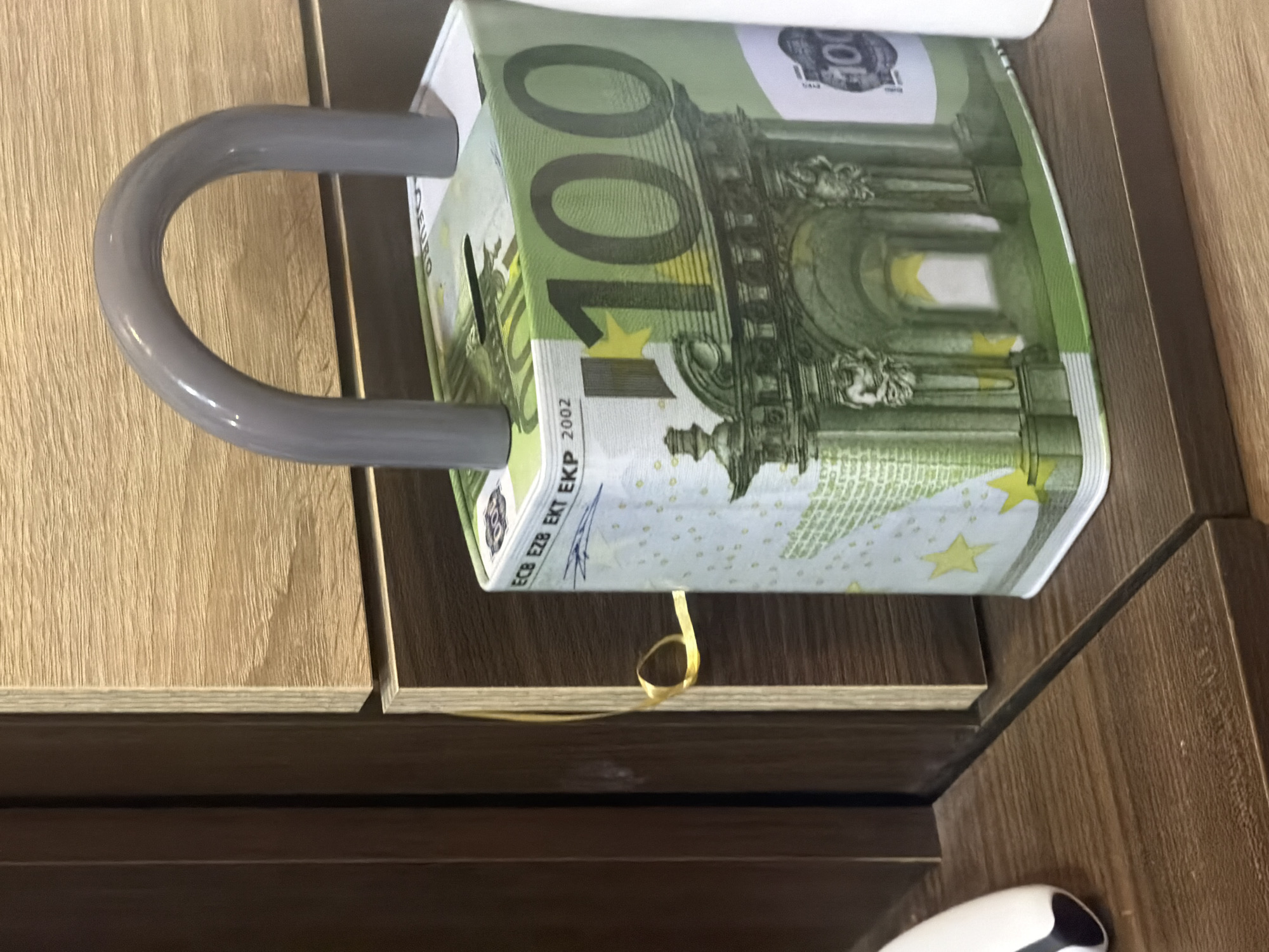 The image displays a roll of paper designed to resemble a 100 Euro banknote, held by a gray, U-shaped dispenser. The roll is horizontally oriented, showcasing the distinctive green and yellow hues of the banknote, with the large number "100" clearly visible, along with architectural details characteristic of Euro currency. Text such as "EURO" and "ECB EZB EKT EKP 2002" can be seen on the paper.
The dispenser itself is mounted on or integrated into a light-colored wooden surface, which exhibits a vertical grain pattern. To the right of this, a darker, possibly walnut-toned wooden surface is visible, suggesting a piece of furniture or wall paneling. A thin, golden, loop-shaped ribbon or string dangles from the underside of the banknote roll.
In the bottom right corner of the original image, a partial view of a white, sleek object with a black section is visible, possibly an electronic device like a computer mouse. The overall scene suggests an indoor setting, potentially a novelty item in a home or office environment rather than a typical bathroom. No people are present, and no specific time of day or weather conditions can be determined.