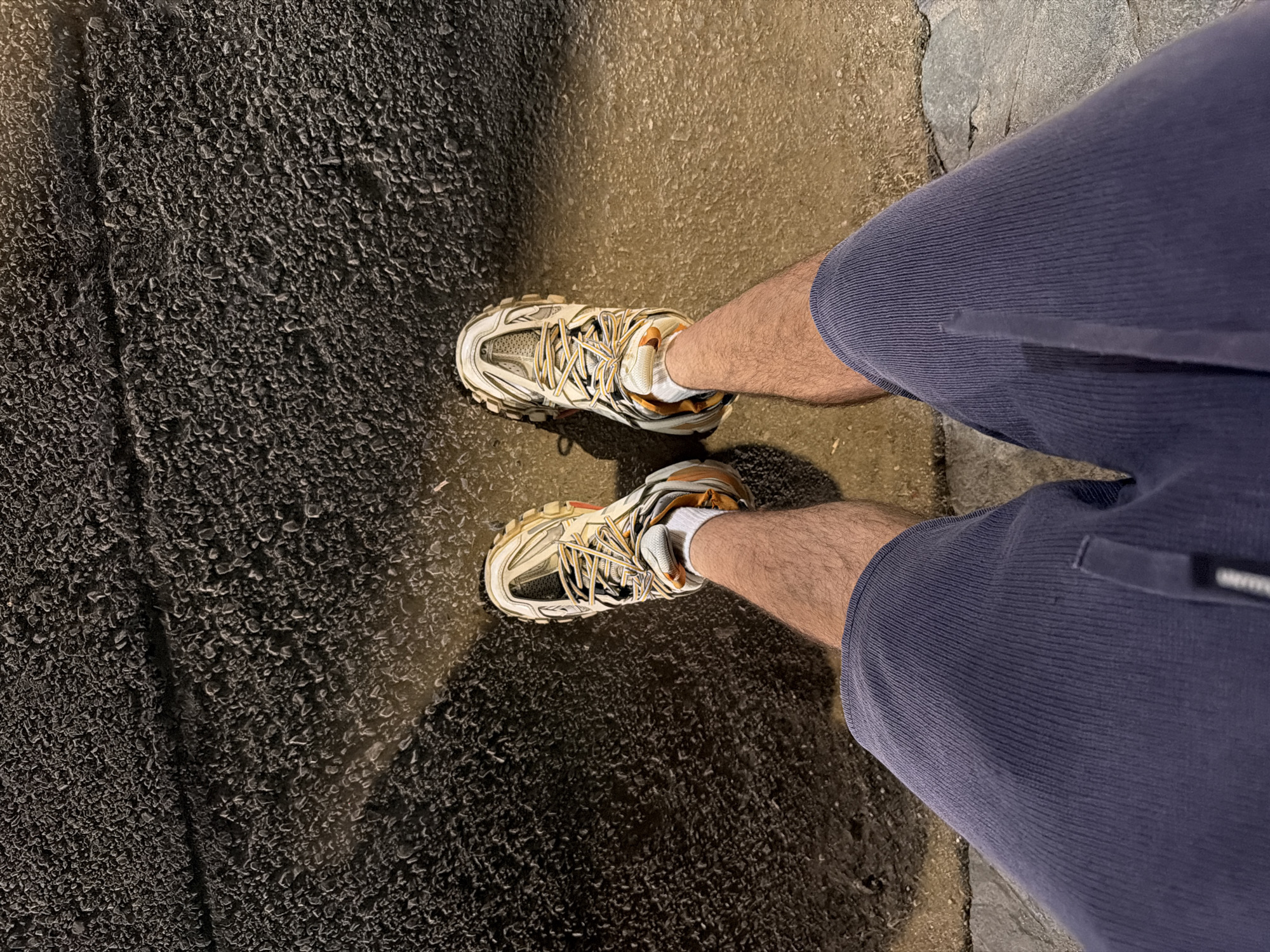 The image presents a first-person, top-down view of an individual's lower legs and feet. The specific location within Sofia, Bulgaria, cannot be determined from the visual information provided. The subject is wearing dark blue, vertically ribbed corduroy shorts, white ankle socks, and multi-paneled athletic shoes. The footwear is predominantly off-white and gray with orange accents, featuring two-tone laces and robust, textured soles. The individual stands on a textured ground surface. The upper portion of the ground exhibits a dark, heavily granular texture consistent with coarse asphalt. This surface transitions to a lighter, somewhat smoother, mottled ground directly beneath and around the feet, separated by a distinct linear seam or crack. A bright, directional light source, positioned behind the viewer, illuminates the scene, casting forward shadows from the feet and creating a localized warm glow on the ground immediately surrounding the footwear.