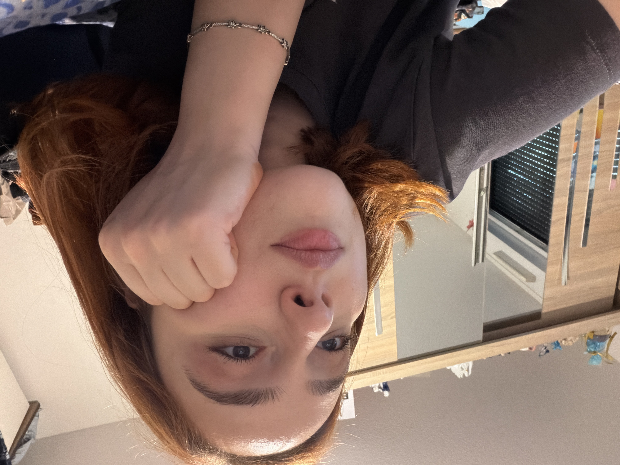 In a room with light-colored walls and a light wood sliding door closet, a young woman with auburn hair rests her head on her hand. She wears a dark grey t-shirt and a bracelet with small star-shaped charms. The closet doors have vertical metal handles. On top of the closet, several small decorative objects are visible, including a blue and yellow wrapped item and white figurines. The overall lighting suggests an indoor setting, likely a bedroom, with a slightly cool tone. There is no indication of the location being in Strasbourg, France.