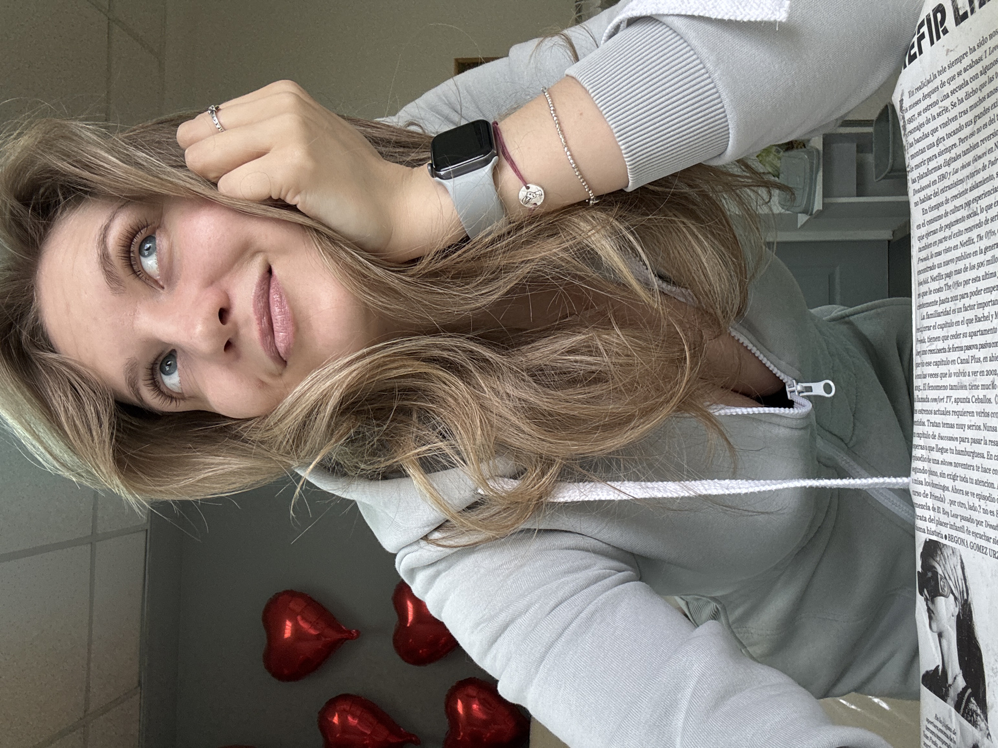 A woman with long blonde hair, wearing a grey hoodie, looks up and to the left. She is wearing a ring, a watch, and bracelets on her left arm. Red heart-shaped balloons are visible in the background, near a gray wall. A newspaper-like publication is nearby. The location is not discernible as being in Izhevsk, Russia, based on the visible details.