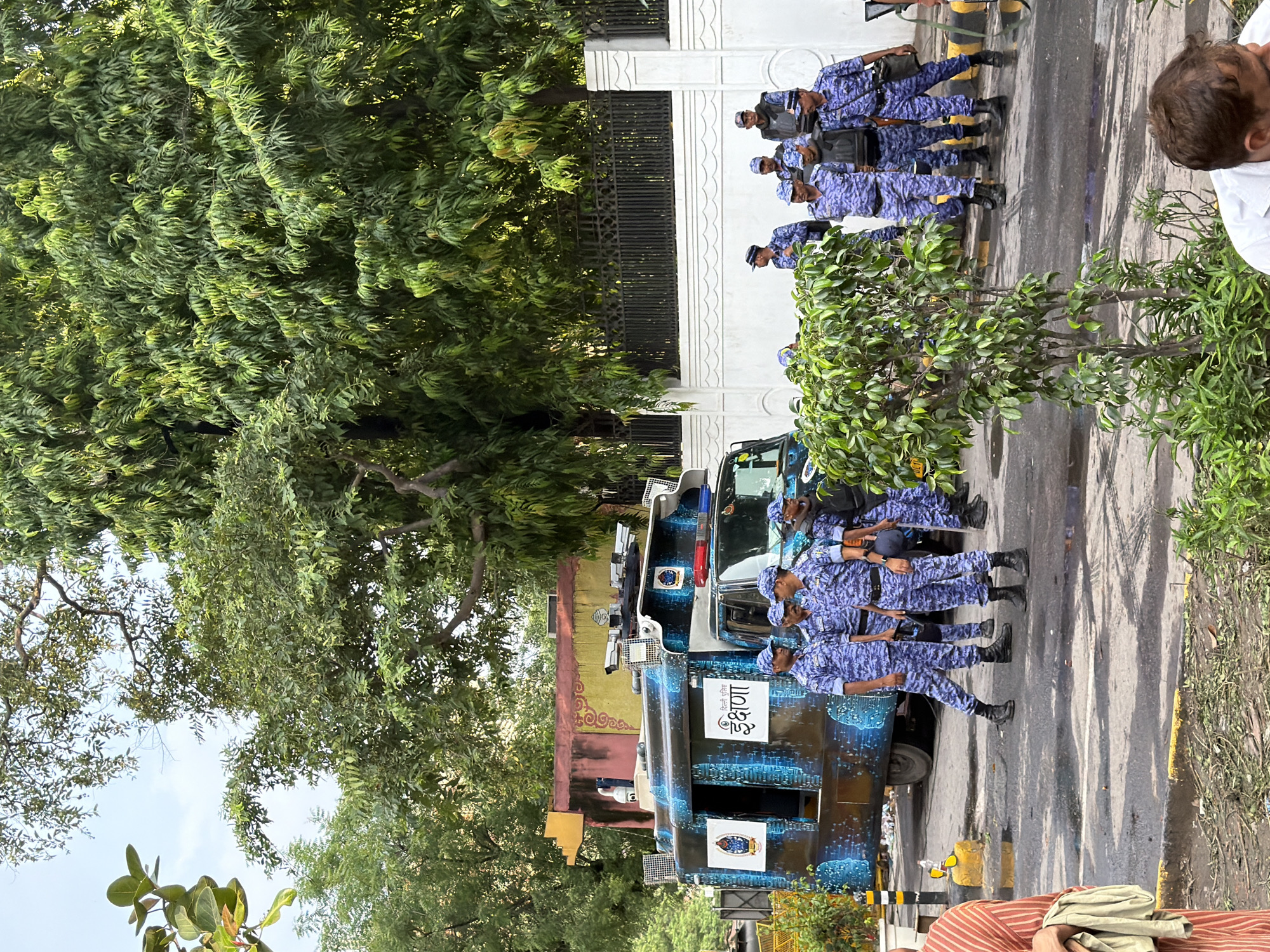 The image depicts a scene on a street in New Delhi, India, with a specialized police van and several officers present. The van, adorned with a blue, galaxy-like pattern and the word "ईक्षणा" (Ikshana) in white lettering, appears to be a surveillance or mobile command unit. It is equipped with multiple cameras and lights on its roof.
A group of police officers, all wearing matching blue camouflage uniforms and caps, stand near the van. Some are positioned in front of it, while others are further back. One officer in the foreground is looking away from the camera. A person wearing a striped orange and white garment is partially visible on the left side of the frame, holding a light-colored cloth.
The environment includes lush green trees in the background, a white wall with decorative molding, and a black metal fence. The ground is paved and appears wet in places, with fallen leaves and debris scattered around. The overall impression is one of security and official activity.