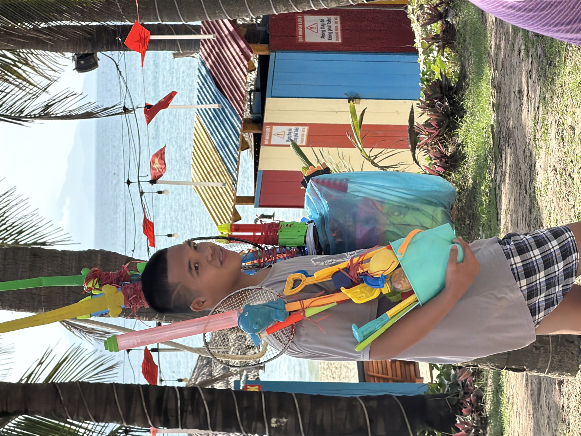 A young man stands at a beach location near Đà Nẵng, Vietnam, holding a blue beach bucket filled with toys. He also has badminton rackets. Behind him is a blue plastic bag containing various items, possibly water toys or beach gear. In the background, the beach and ocean are visible, as well as a structure with colorful stripes and a sign in Vietnamese. Palm trees frame the scene, and there are small red flags strung up above. The area seems to be a tourist or recreational spot with a casual, outdoor atmosphere.