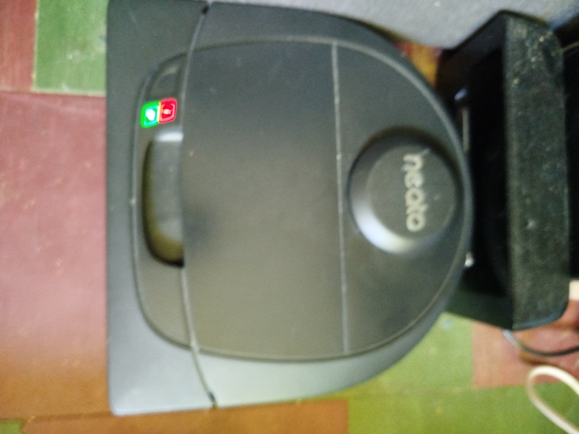 The image shows a Neato robotic vacuum cleaner in its charging base. The vacuum cleaner is black and has a distinctive D-shape. It is sitting on a floor with a pattern of green and red tiles. The charging base is also black and has a cable connected to it. The background is blurred, but it appears to be an indoor setting. The image is taken from a top-down perspective. There are no people visible in the image. The location in the city of Pavlohrad, Ukraine is not specified.
