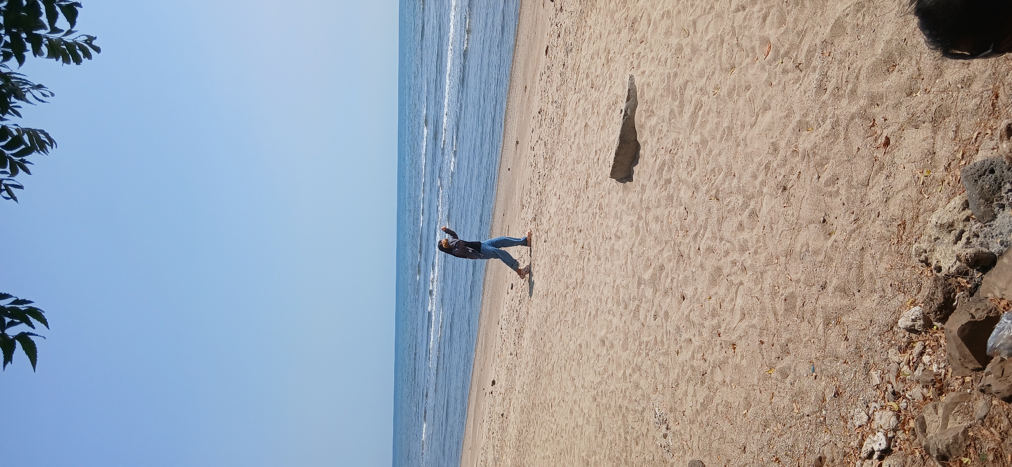 A medium shot shows a woman walking on a sandy beach in Banyuglugur, Indonesia, with the ocean in the background. The sky is clear and blue. The woman, who is wearing a dark jacket, a headscarf, and blue jeans, is in the middle of the frame. She appears to be holding something yellow up to her face with her right hand, possibly a phone or camera. She is walking from left to right. The beach is covered in light brown sand with some scattered leaves and debris. A concrete object is visible on the sand to the right of the woman. In the foreground, rocks and leaves are scattered on the ground.