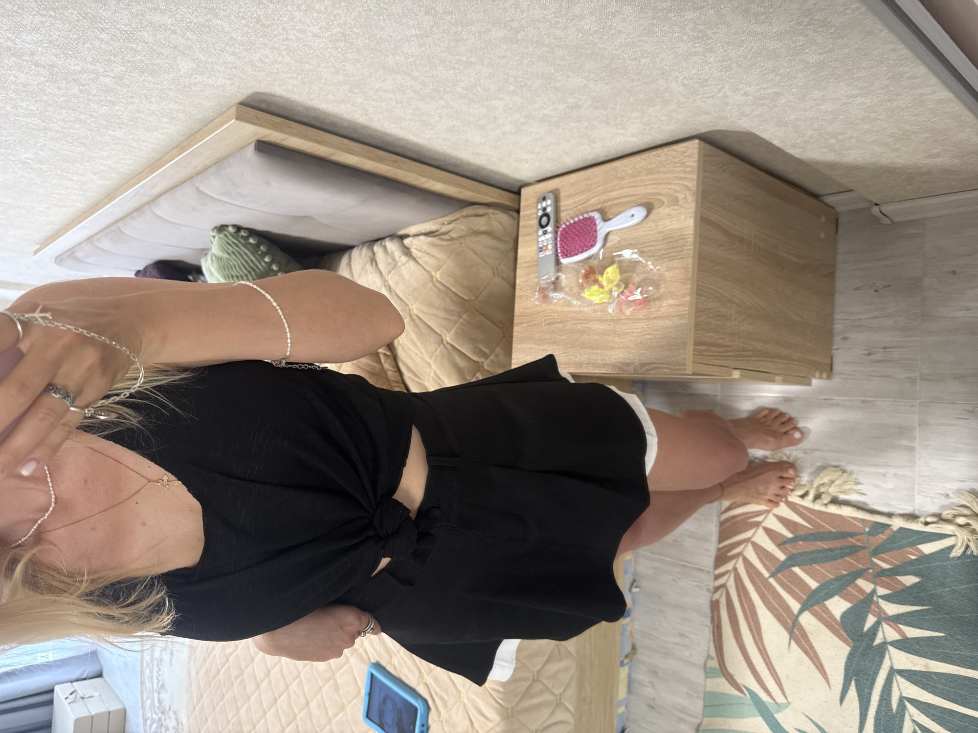 A woman is standing in a bedroom, facing a mirror. She is wearing a black crop top and a black skirt with a white underskirt. The woman has blonde hair and is wearing jewelry on her neck and wrists. To her right, there is a nightstand with a hairbrush, a remote control, and some candy on it. The room has a bed with a beige headboard and a patterned rug on the floor. The overall lighting is natural and soft.