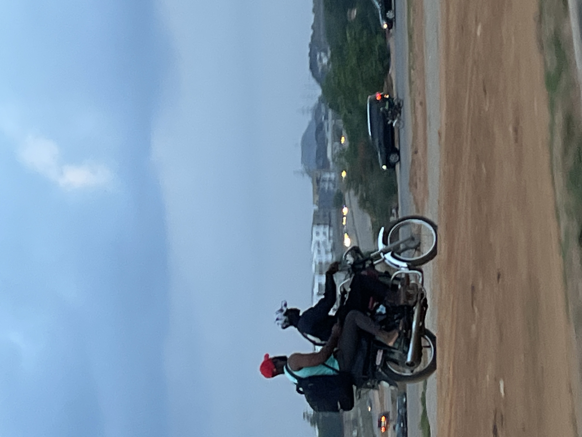 Two men are on a motorcycle traveling on a paved road in Galadimawa Village, Nigeria. One man is wearing a red hat and a light blue tank top. The other man is wearing a helmet and a black jacket. There are cars on the side of the road and buildings in the background. The sky is overcast. The road is bordered by dirt.