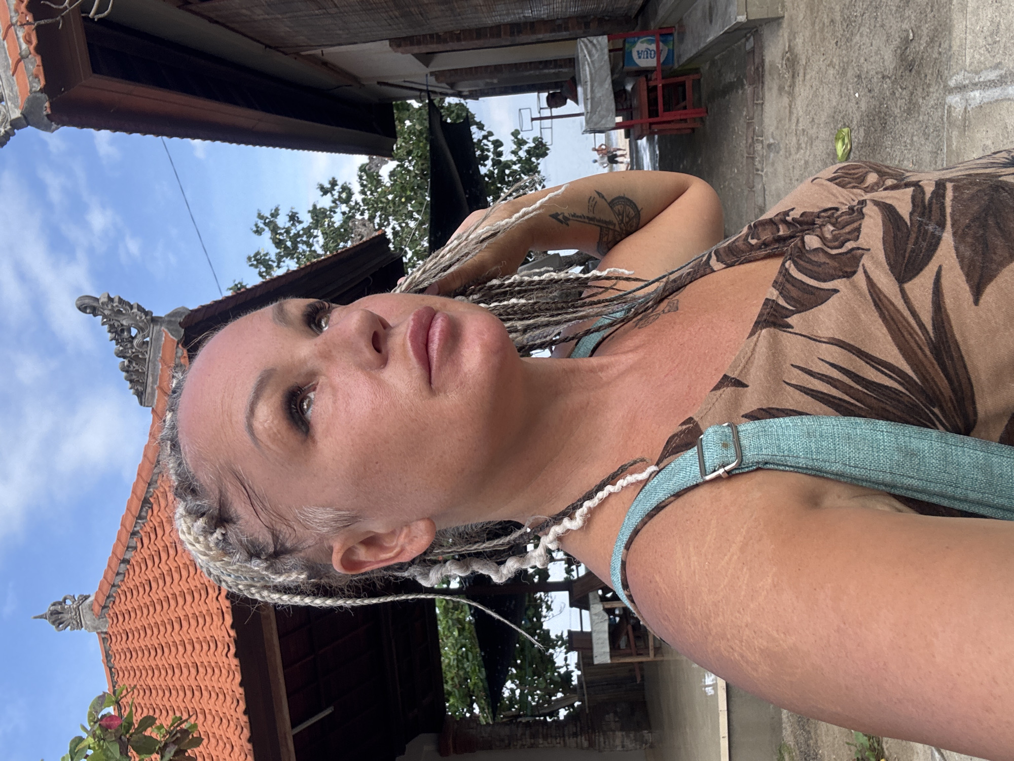 A woman is facing upward in an outdoor setting. She has light skin, light-colored braided hair, and appears to be wearing a printed top with a green strap or harness over her shoulder. Her left arm is raised and shows a tattoo.
The location features architecture characteristic of Bali, Indonesia. A building with a traditional Balinese roof is visible on the left side of the frame. In the background, there are structures that look like small shops or stalls.
The location can be identified as Kuta Selatan, Indonesia based on the distinctive Balinese architectural style. The weather appears to be sunny and clear.