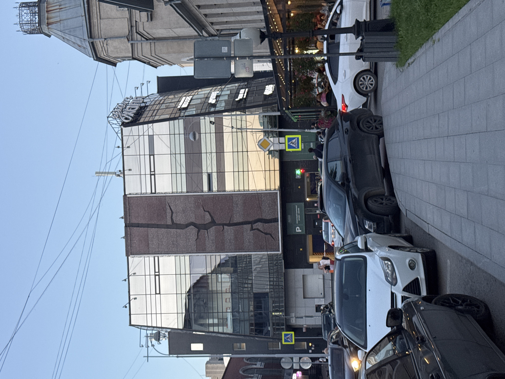 The image depicts a street scene in Yekaterinburg, Russia, likely during the evening. The foreground shows a sidewalk with gray paving stones and a patch of green grass. Several cars are parked or moving on the street, including a white Ford Focus and a black SUV. In the middle ground, a modern building with a glass facade and a brick section featuring a tree-like graphic dominates the view. The building has signs that are partially visible, including "DOORS" and "SNOW" in English. To the right, an older, ornate tower with a weathered metal cap stands next to the modern building. A covered outdoor seating area with patrons is visible near the base of the modern building. Traffic lights and pedestrian crossing signs are also present. The sky is a clear, pale blue, indicating dusk or early evening.