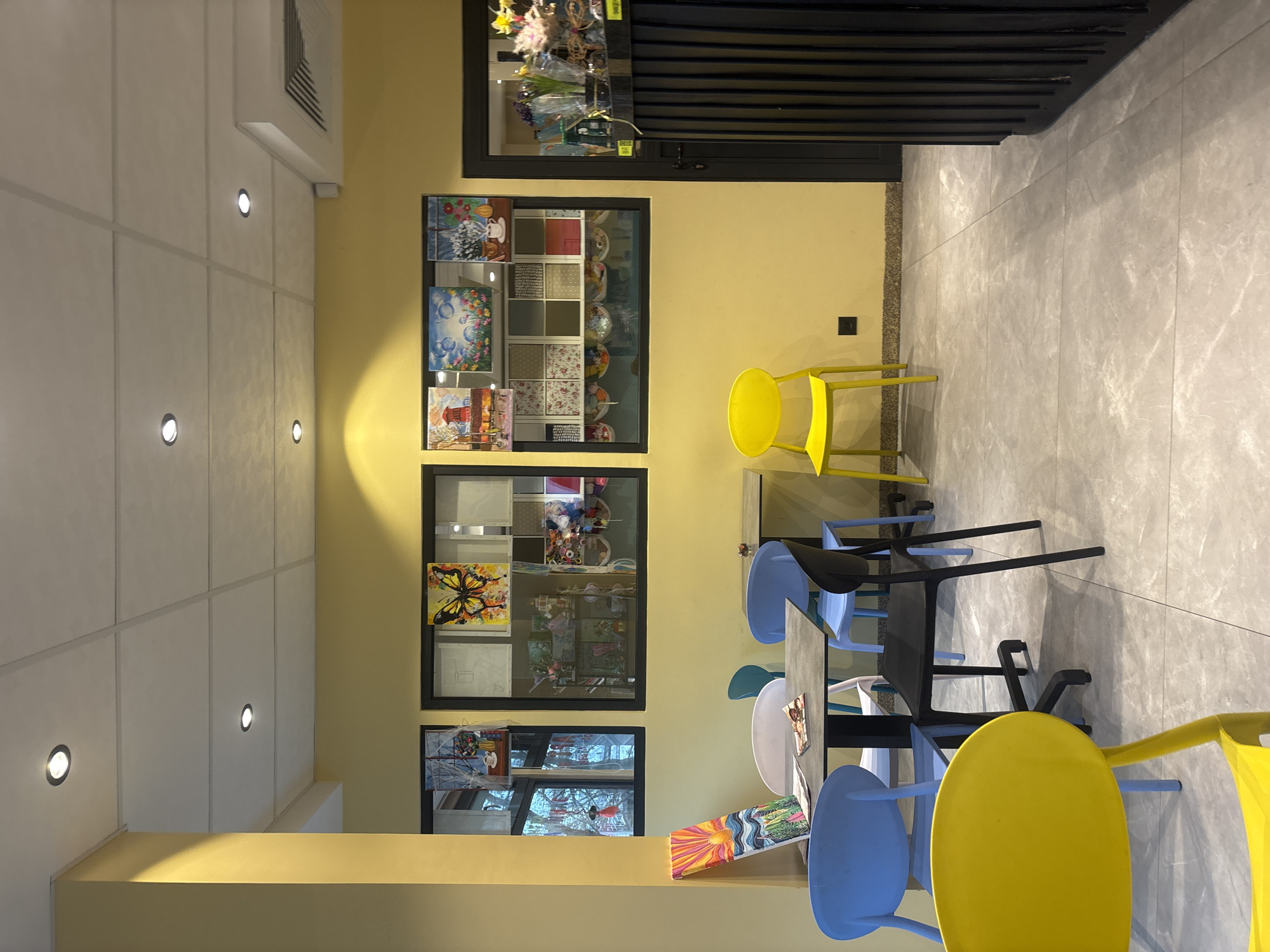 The image shows an interior space with a yellow wall, white ceiling with recessed lighting, and a grey marble-patterned floor. Three framed sets of images or artwork are hung on the yellow wall. One frame appears to have a grid of smaller images, while others contain what seem to be paintings. In the foreground are several chairs, mostly yellow and light blue, around a table. The scene seems to be in a cafe or restaurant setting. There is no indication of this being in Burgas, Bulgaria, but the location may be in an Eastern European city. No people are visible. The environment is modern and well-lit.