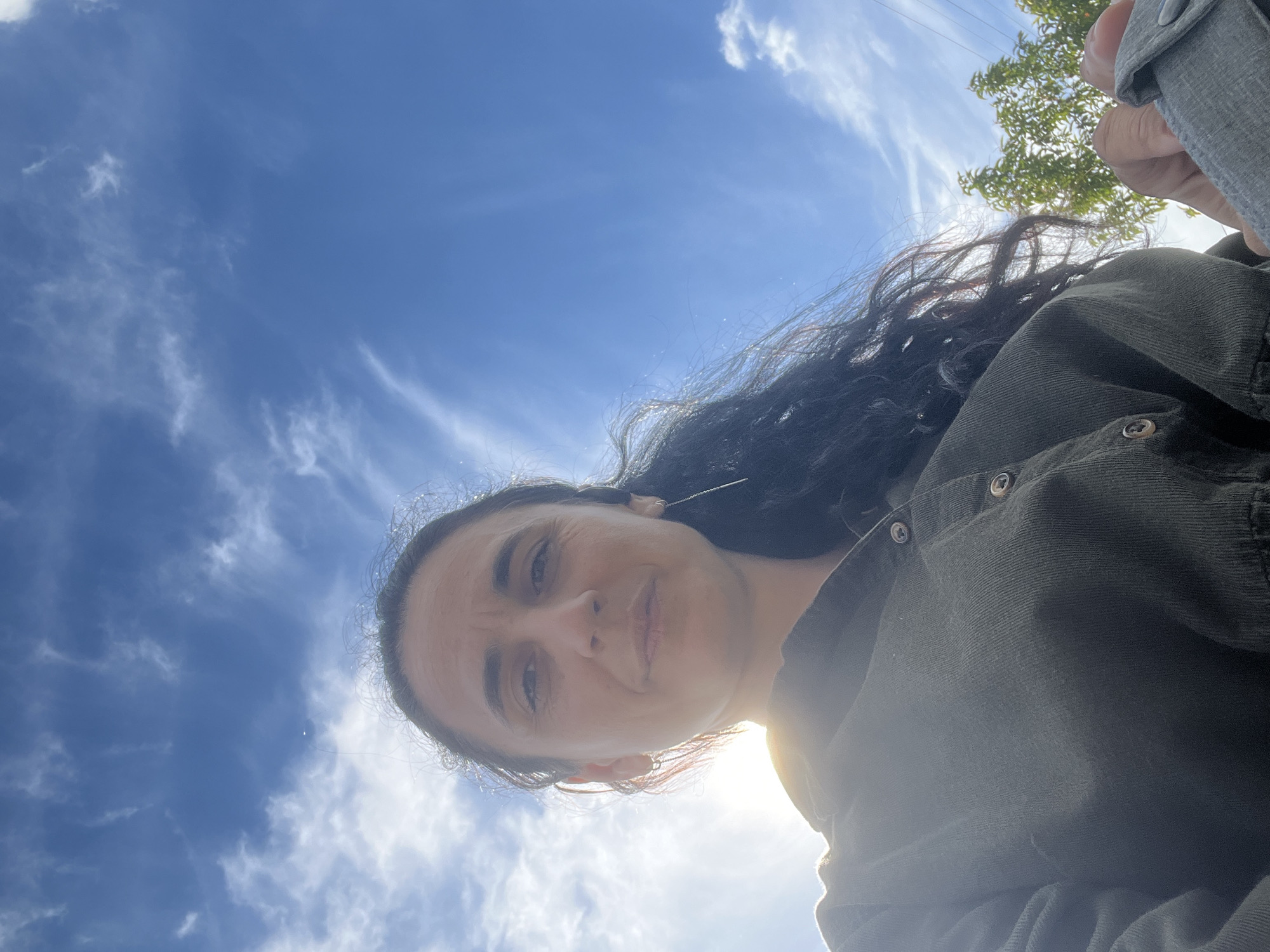 A low-angle, outdoor, selfie-style shot depicts a woman with long, dark, curly hair against a bright blue sky with wispy white clouds. She is looking directly at the camera with a slight smile. The sun is positioned to her left, creating a lens flare and illuminating her face. She is wearing a dark, button-down shirt. A thin, dangling earring is visible in her right ear. In the background, out of focus, are green trees and foliage. The image appears to be taken in the village of Onokivtsi, Ukraine, given the context provided.