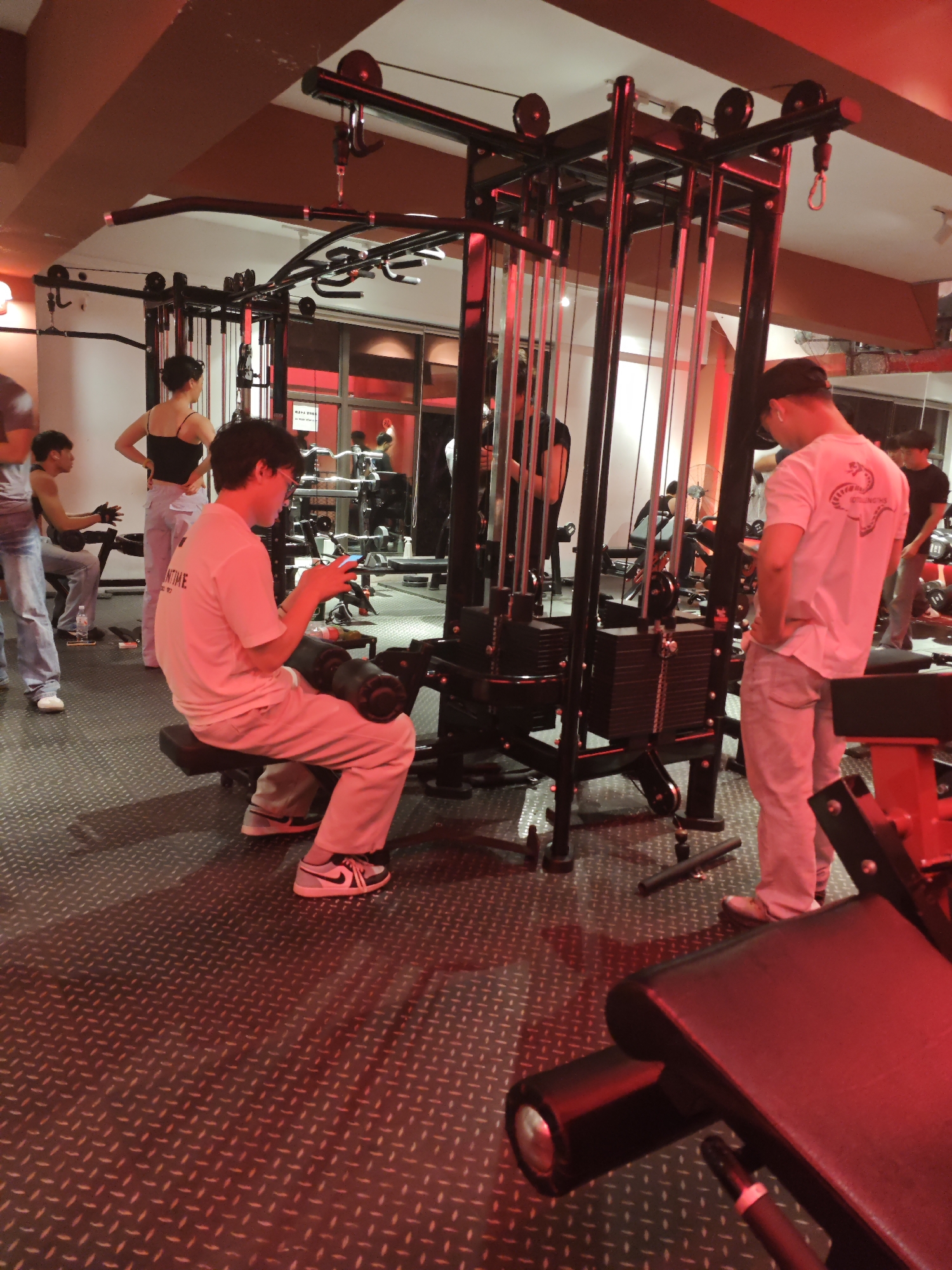 The image depicts an indoor gym scene in Chongqing High-tech Zone, China, illuminated by prominent red artificial lighting. Several individuals are present, engaged in or resting from exercise.
In the foreground, a man wearing glasses, a white t-shirt (with partially visible text like "NTIME" or "ONE TIME"), and light-colored pants sits on a leg curl machine, engrossed in his mobile phone. His black and white sneakers are visible. To his right, another man, wearing a white t-shirt with a graphic of a coiled snake and illegible text below it, stands near a large black cable crossover machine with visible weight stacks, looking downwards.
Behind these individuals, a woman in a black tank top and light-colored pants stands with her back to the viewer, hands on her hips. Further in the background, other gym-goers and various pieces of exercise equipment are visible through glass partitions and within the main area. A water bottle is on the floor near a leg press machine where another person is seated in the mid-ground.
The gym floor is dark with a distinct textured, diamond-plate pattern. The pervasive red lighting creates a dramatic and warm ambiance, casting red reflections on surfaces and equipment. A sign visible through a glass partition in the background is highly reflective, showing some English text that appears to be "PLAYA PARADISE" and possibly some Korean characters, but it is largely unreadable. The scene suggests an active gym environment during evening hours or within a dedicated, mood-lit space.