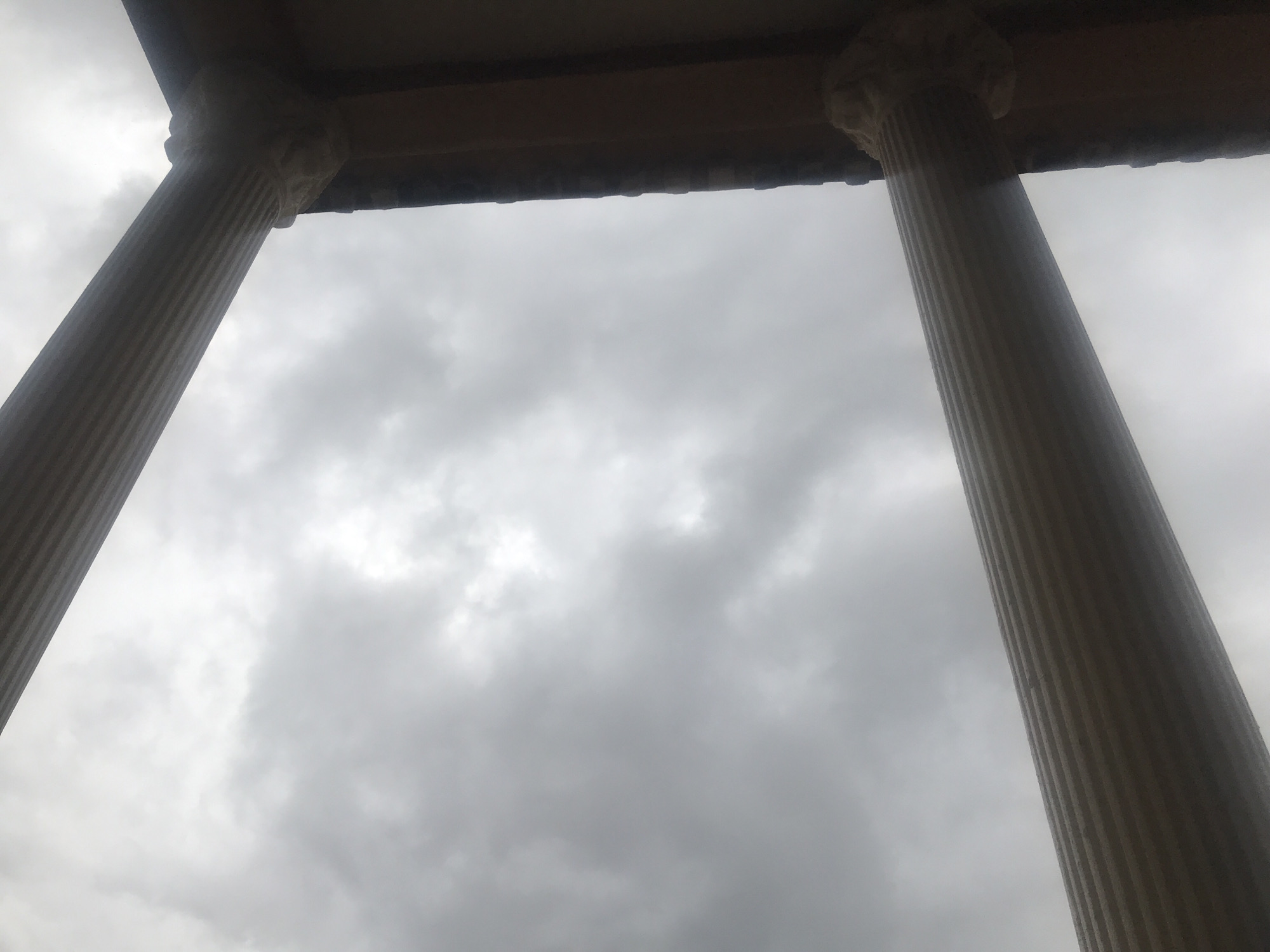 The image shows a low-angle shot looking up at two fluted columns supporting an overhang. The sky above is overcast with dense gray clouds. The columns appear to be made of stone or concrete and are ribbed vertically. The architectural style suggests a classical or neoclassical design. The location is identified as New Stadium, Nigeria. There are no people or visible actions captured in this image.