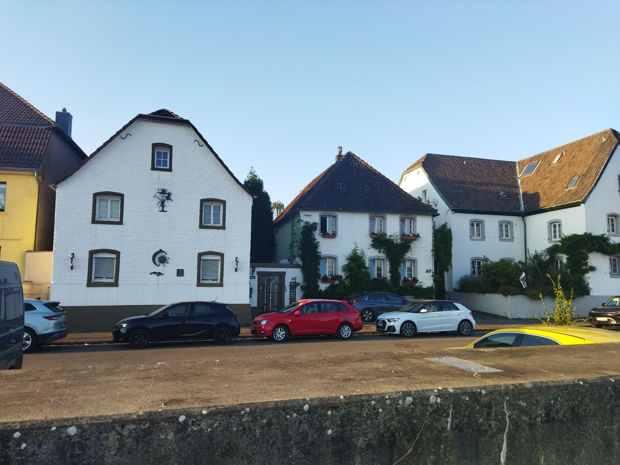 The image shows a row of white buildings with tiled roofs, characteristic of older European architecture, lining a street in Leverkusen, Germany. The buildings have numerous windows and some are adorned with ivy and flower boxes, suggesting a residential area. Several cars are parked along the street. From left to right, a dark gray van, a light blue SUV, a black hatchback, a red station wagon, a dark gray sedan, a white hatchback, and a dark brown sedan are visible. The sky is clear and blue, indicating daytime. The street appears to be paved, and there is a concrete wall in the foreground, suggesting a slight elevation change. The overall scene is peaceful and orderly.