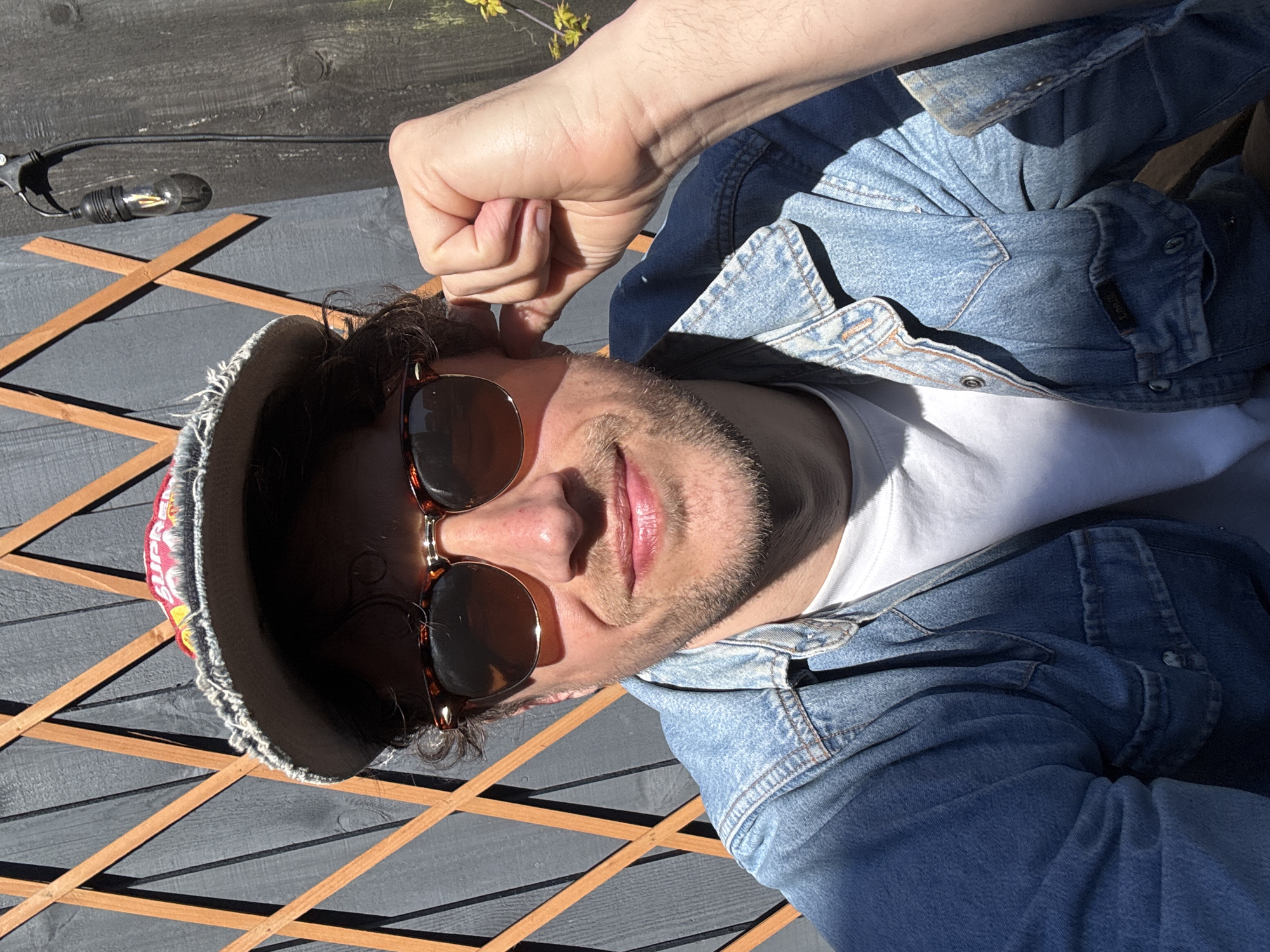 A person is shown from the waist up, wearing a white t-shirt and denim jacket, with sunglasses and a brimmed hat. They are positioned on an outdoor, wooden, gray-painted structure, with tan-colored lattice details. A string of lights is visible in the upper left corner of the scene. The location is not identifiable in this image.
