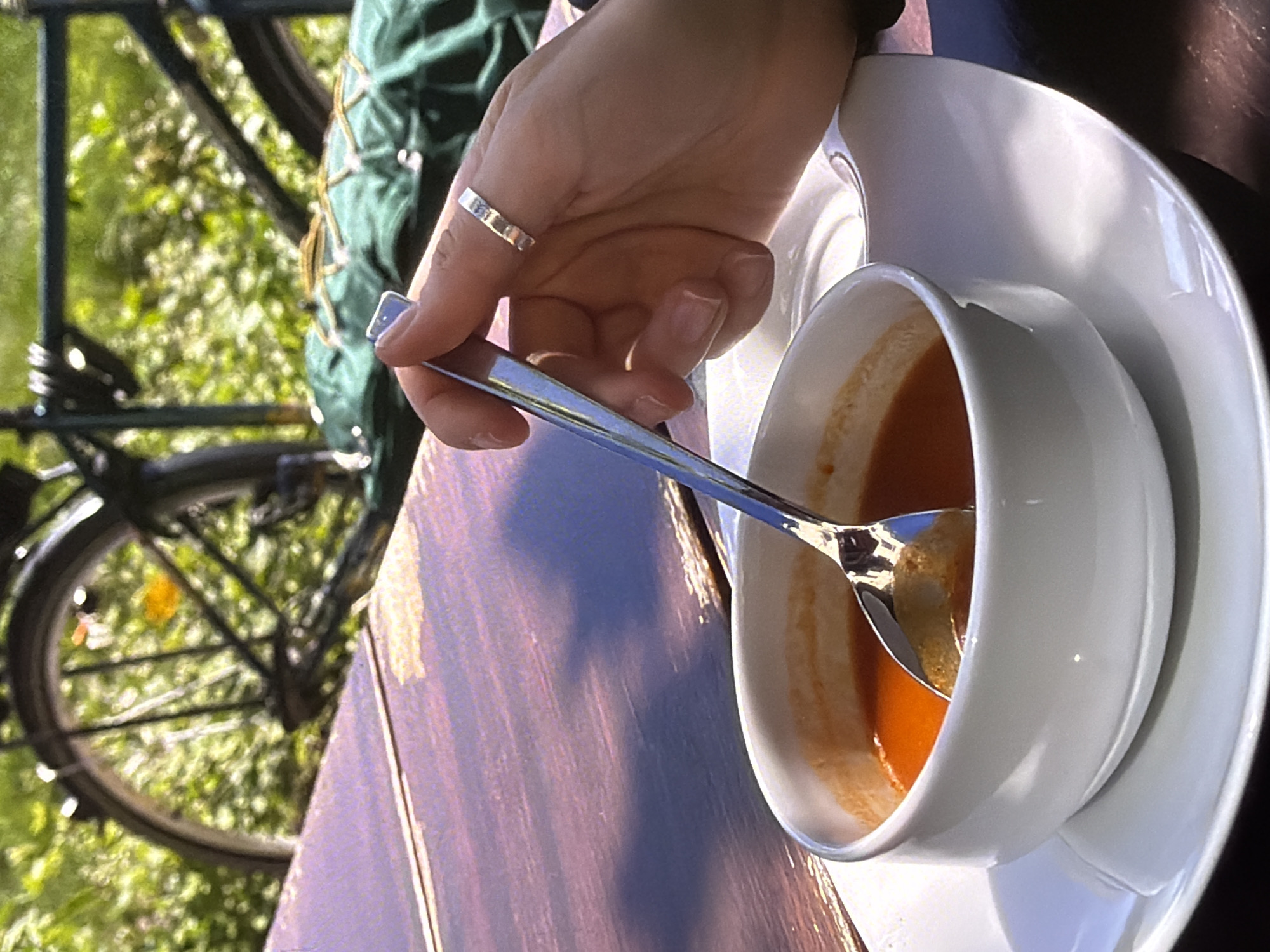 A hand wearing a silver ring is poised over a white bowl of tomato soup. A silver spoon is submerged in the soup, with a bit of solid material visible on its surface. The soup is a vibrant orange color and there are some remnants of soup clinging to the sides of the bowl. The bowl is resting on a white saucer. The scene appears to be outdoors, with a blurred background of green foliage and a bicycle wheel. The table surface beneath the bowl and saucer is a warm brown color with visible wood grain. The lighting suggests it is daytime, possibly late afternoon given the warm tones. The overall mood is casual and relaxed. There is no visible text in the image.