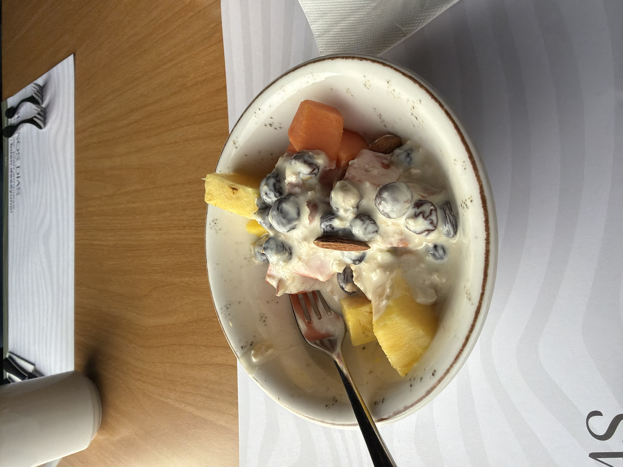 A close-up, eye-level shot shows a white ceramic bowl filled with a fruit salad. The salad contains chunks of pineapple, papaya, blueberries, and what appears to be a sliver of almond. The fruit is coated in a creamy white dressing, possibly yogurt or whipped cream, and there are some visible specks of dark color, likely from the blueberries, on the rim of the bowl. A silver fork is partially submerged in the salad. The bowl rests on a white placemat with a subtle wave pattern. In the background, a wooden table is visible, and on the far left, the bottom of a white mug or pitcher is present. Two black forks are placed on a white paper mat with Spanish text that reads "¡BUEN DÍA! SON DIAS." The image is likely taken indoors, possibly at a breakfast establishment in Bogotá, Colombia.
