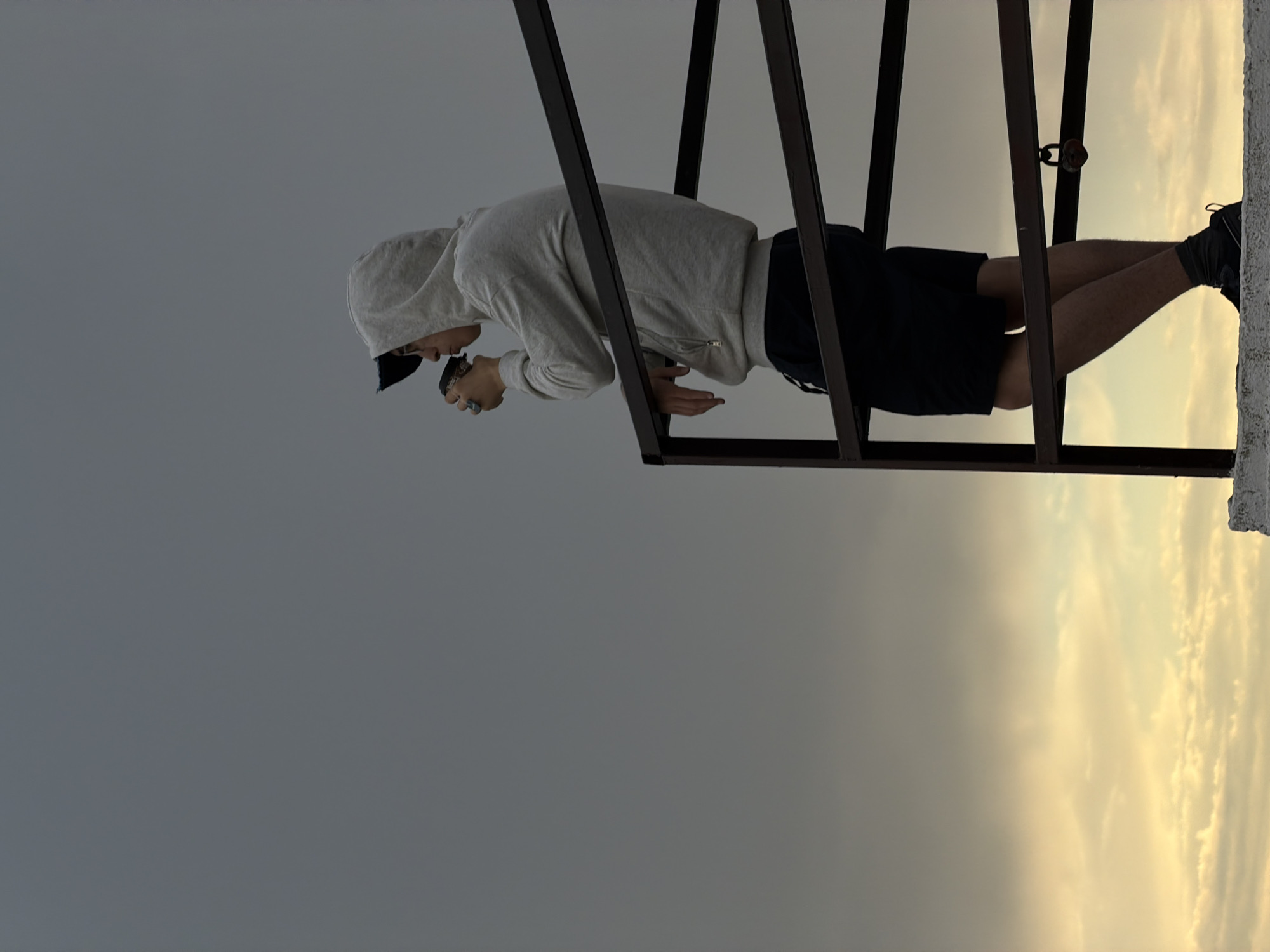 A young person in a gray hooded sweatshirt and black shorts stands on a concrete ledge, holding a dark cup to their mouth as if drinking. They are leaning against a dark metal railing with their left arm. A black baseball cap is worn backward under the hood. The background is a vast, overcast sky with lighter clouds near the horizon. A rusted padlock hangs from the railing. The setting appears to be an elevated outdoor location, possibly a rooftop or observation deck, with a view of the sky at dusk. The location is Goryachevodsky, Russia.