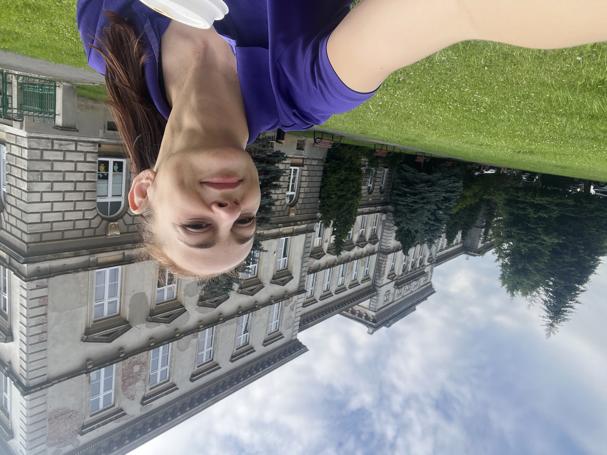 A woman in a purple shirt is taking a selfie outdoors in front of a large, old building. The building appears to be made of stone and has many windows. There are trees and a grassy area in front of the building. The sky is cloudy. The woman is smiling and holding a white cup in her hand.