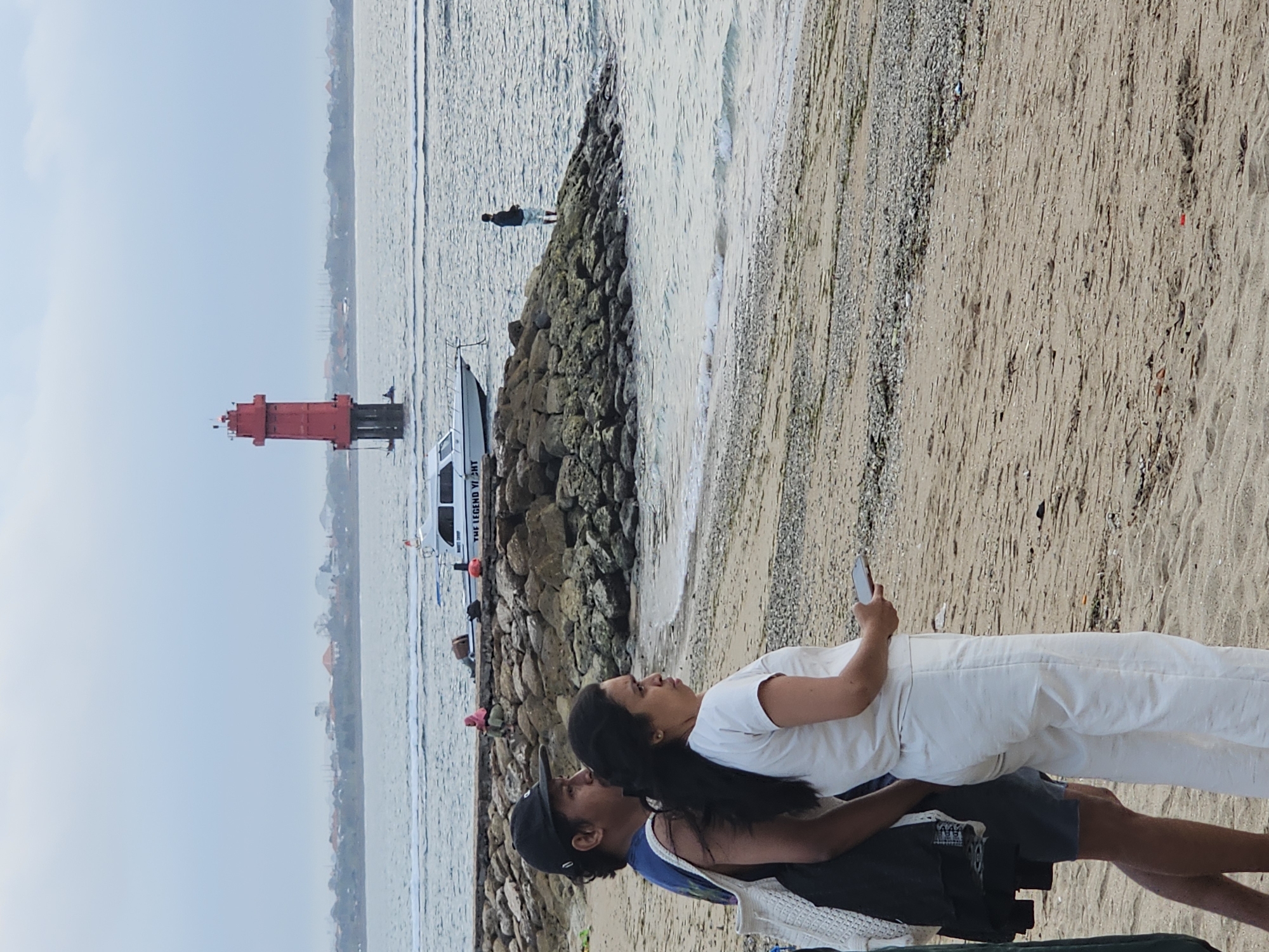 The image shows a beach scene in Denpasar, Indonesia. In the foreground, two individuals, a man and a woman, stand on the sand facing away from the camera, looking out at the water. The man is wearing a dark blue tank top and a black baseball cap. The woman is wearing a white top and white pants and holding a mobile phone. Behind them, a breakwater made of large rocks separates the sandy beach from the sea. In the middle ground, several boats are visible, including a white speedboat with "THE LEGEND YACHT" written on its side. Further in the distance, a tall, red lighthouse stands in the water. The sky is overcast. The overall impression is a calm, overcast day at a coastal area.