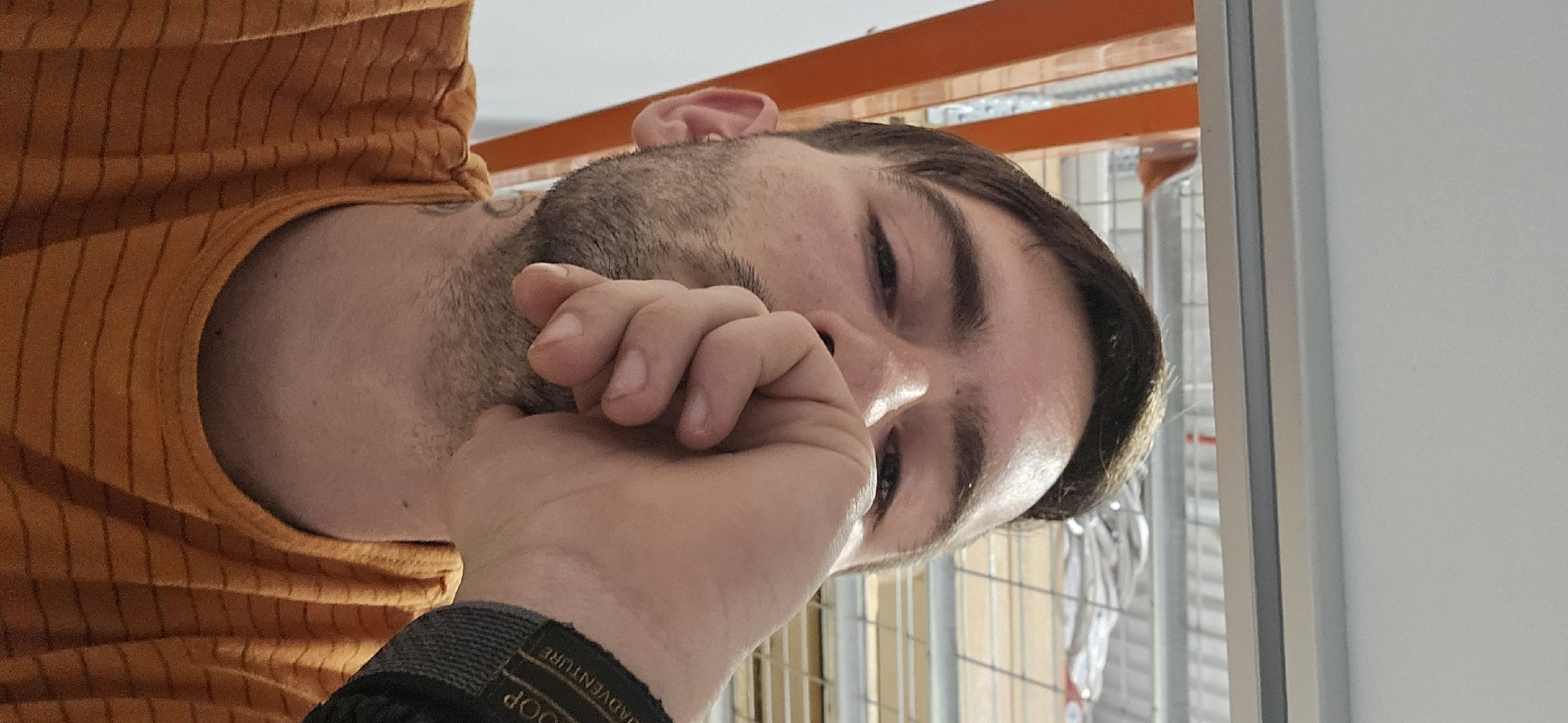 The image, rotated 90 degrees clockwise, presents a close-up, low-angle portrait of a young man. He is shown from the chest up, wearing an orange t-shirt with vertical stripes. His left hand is raised, with fingers loosely curled, positioned near his mouth and nose. On his left wrist, a dark fabric wristband or watch strap is visible, bearing the text "LOOP ADVENTURE".
The man has dark hair, styled shorter on the sides and slightly longer on top, with prominent dark eyebrows and stubble or a short beard. His eyes are open, looking slightly upwards and to the right, conveying a neutral to mildly pensive expression.
The background suggests an indoor, potentially industrial or commercial environment. Visible behind him are orange metal beams or structural elements, likely part of shelving or a building's framework. Through these, metal grating or fencing is discernible. To the right of the frame, a plain white wall or vertical surface is present, possibly part of a window or door frame. The bright illumination indicates either ample natural light from windows or strong artificial lighting, suggesting it is daytime. The overall scene portrays a moment of contemplation or a posed shot within a functional, non-residential space.