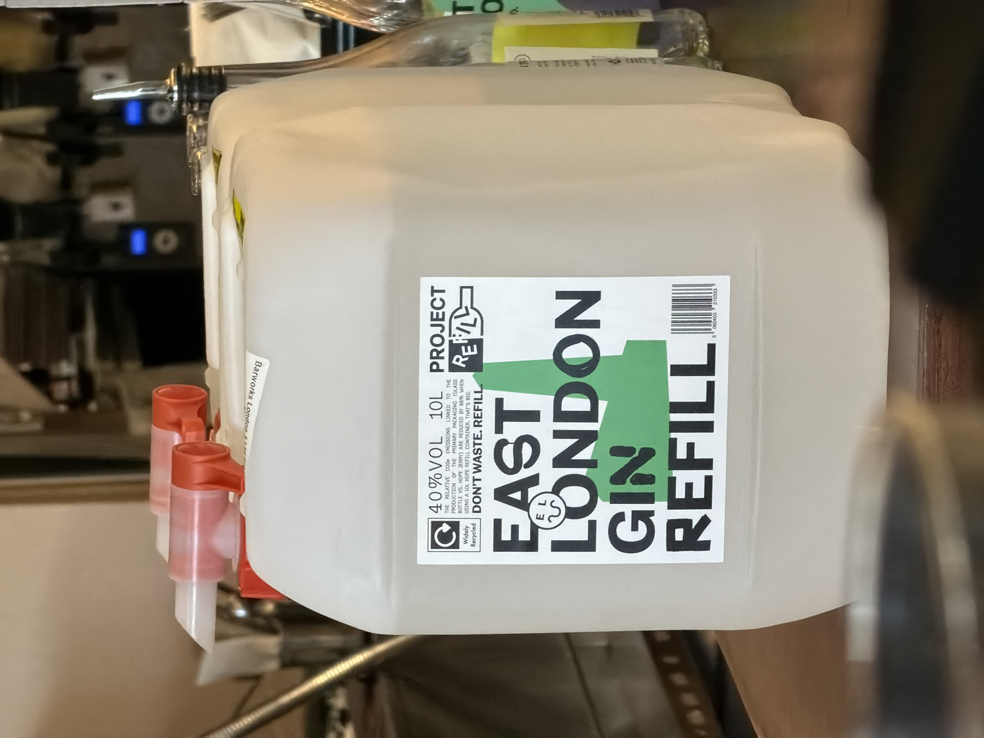 A 10-liter container of East London Gin Refill is the central focus. The container is white and translucent, with a prominent label featuring the brand name in bold, green, and black lettering. The label also includes the text "Project Refill" and information about the reduced CO2 emissions associated with using the refill container. The container has two red dispensing spouts attached to it. The background is blurred, suggesting an indoor bar setting with other bottles and equipment visible.