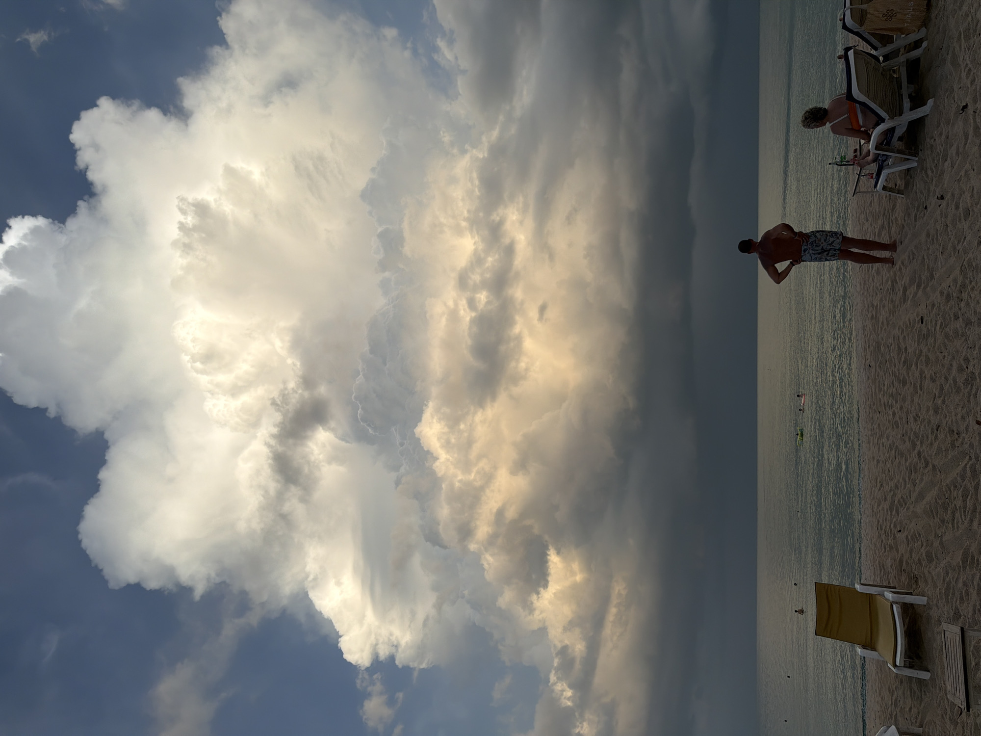 A panoramic shot captures a serene beach scene on Ko Samui, Thailand, under a dramatic sky. Towering cumulonimbus clouds dominate the upper portion of the frame, illuminated by warm, golden light that contrasts with the darker, grayer clouds lower down, suggesting an approaching storm. The sea is calm, with a few people visible in the distance, some in the water and others on inflatable rafts. In the foreground, the sandy beach is sparsely populated. Two beach chairs are occupied: one by a man standing with his hands on his hips, facing the sea, and another by a woman seated, facing away from the camera. The presence of a table and drinks beside the seated woman suggests a leisurely moment. The overall atmosphere is one of impending change, with the beauty of the natural elements creating a striking visual.