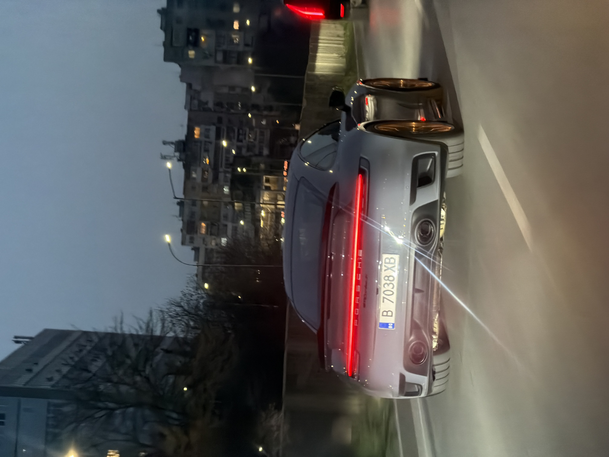 The image depicts a silver Porsche 911 driving on a road at dusk in an urban environment. The car has a red taillight strip spanning its width and a license plate that reads "B 7038 XB". The background features multi-story buildings, possibly apartment complexes, with streetlights illuminating the road. Bare trees are visible along the roadside. The location is not Varna, Bulgaria as the license plate indicates Bucharest, Romania.