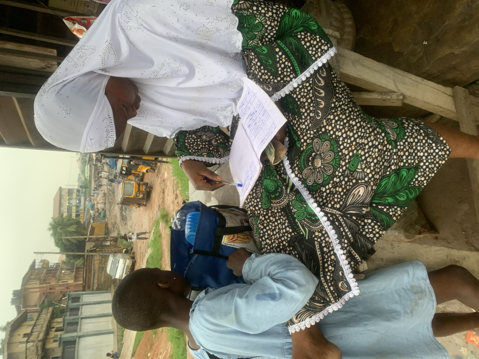 An outdoor scene in Shagamu, Nigeria, depicts two individuals engaged in an activity. On the right, an adult woman wearing a white hijab adorned with rhinestones and a green and black patterned dress is seated. She is holding a pen and writing in an open notebook that rests on her lap. Several currency bills are visible beneath the notebook. To the left, a child in a light blue dress stands facing the woman, looking down at the notebook. A blue bag containing water bottles and other items is placed between them. The background reveals a dirt road with various vehicles, including a yellow auto-rickshaw and a white van, as well as buildings with balconies and corrugated metal structures, characteristic of an urban environment. The overall atmosphere suggests an exchange or educational interaction taking place in a bustling, possibly commercial, area.