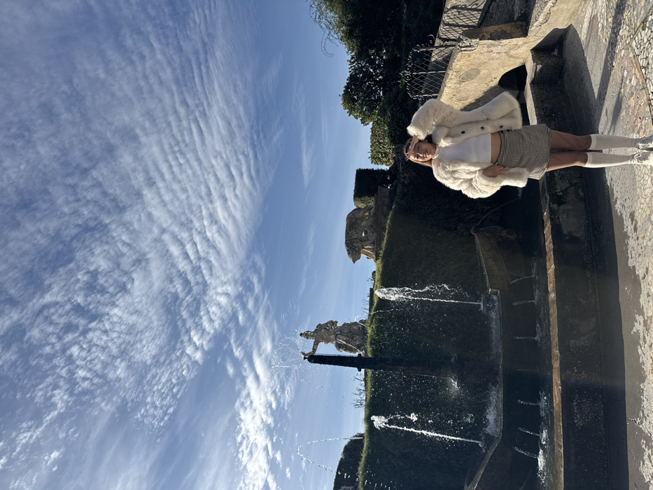 The image captures a sunlit scene within the terraced gardens of the Villa d'Este in Tivoli, Italy. A young female subject stands in the foreground, slightly right of center, facing the viewer with a slight smile. She wears a white fluffy coat over a white top, a gray and white plaid mini-skirt, white knee-high socks, and white sneakers. Her right hand is raised to shade her eyes. To her left, a stone fountain system features a wide lower basin with multiple active water jets, with additional jets flowing from an upper section built into a green, sloped embankment. On an elevated terrace behind the subject and the immediate fountain, two weathered stone statues are visible: a robed classical figure on a pedestal to the left, and a distinct bear statue on the right. Further right, a light-colored stone archway and dark metal railings are present. The background features a clear blue sky with extensive, rippled altocumulus cloud formations.