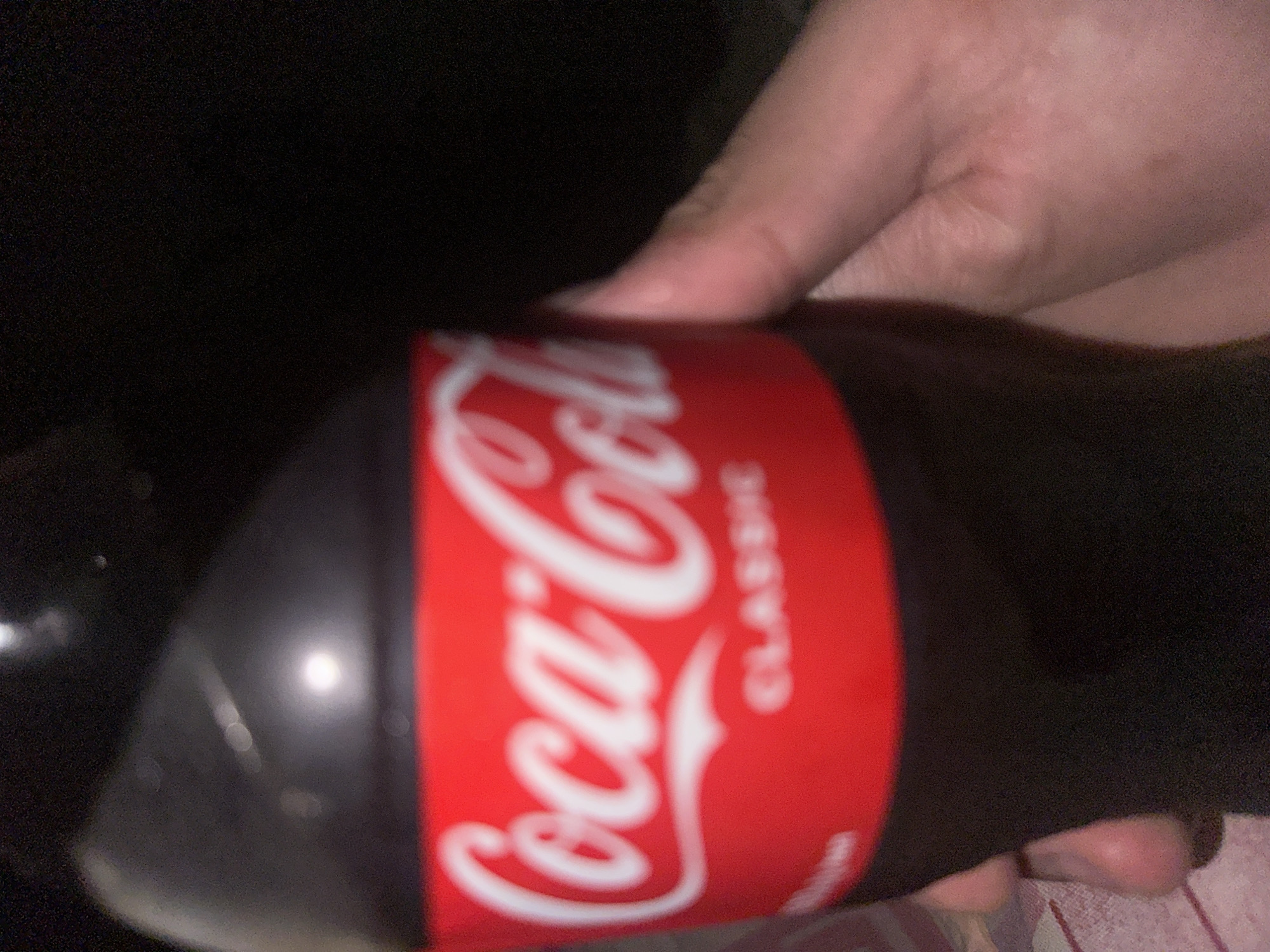 A hand holds a bottle of Coca-Cola Classic. The background is dark and out of focus. The location is Unknown, Unknown.