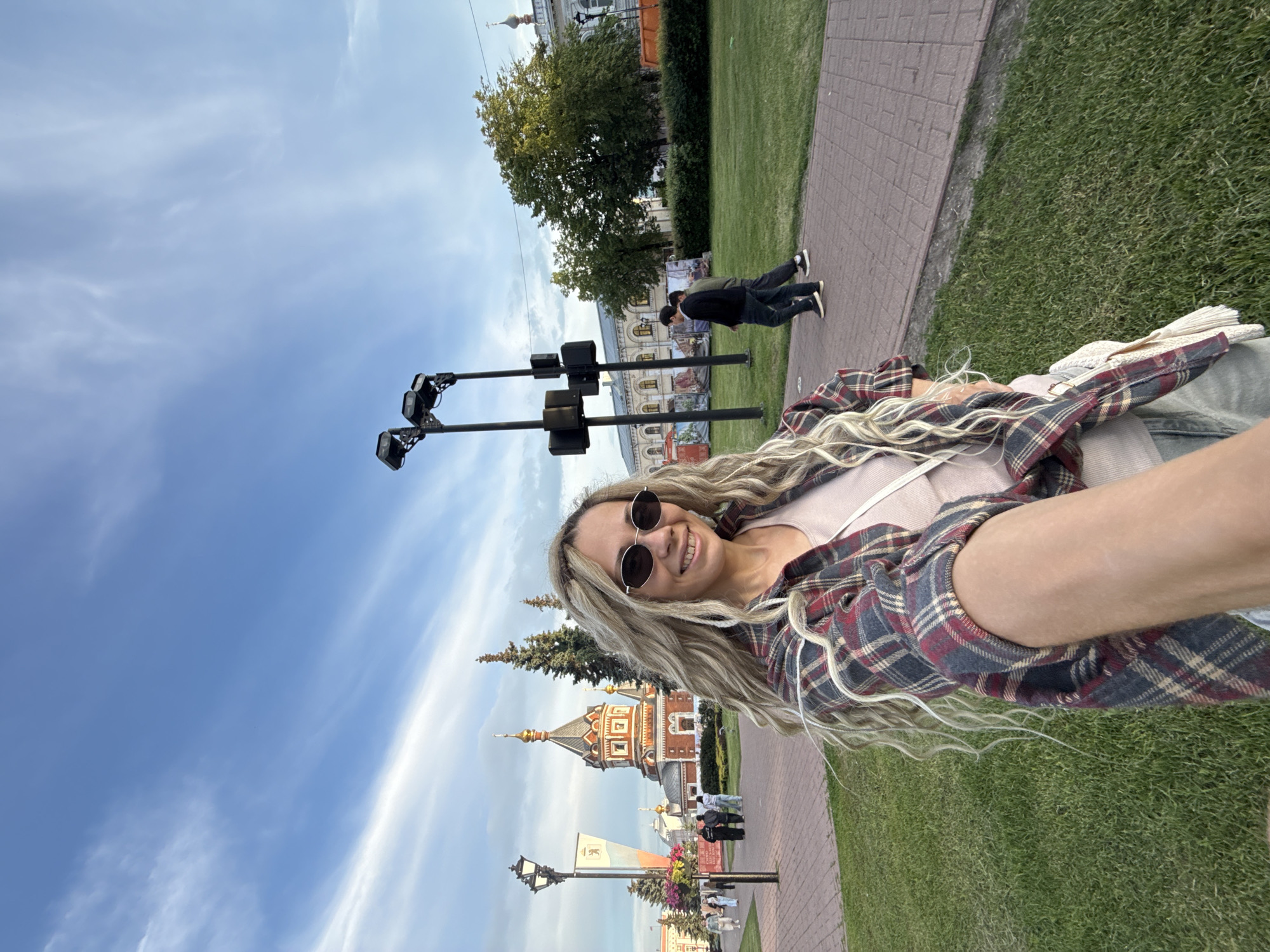 A selfie-style photo, rotated 90 degrees clockwise, captures a smiling woman outdoors. She is in the foreground, looking directly at the camera, wearing dark, round sunglasses and a red, black, and beige plaid flannel shirt over a light-colored top. Her long, wavy blonde hair flows over her shoulders.
Behind her, the scene unfolds under a blue sky with scattered white clouds, suggesting a pleasant daytime setting, possibly late afternoon. To her left, two prominent black poles with multiple light fixtures or cameras are visible. In the mid-ground, a grand architectural building with red brick, white trim, and an ornate, possibly golden-domed or spired roof stands out, suggesting a church or historic structure. Lush green grass lines a paved pathway made of reddish bricks. Several other people are visible in the background, some walking, including two men in dark clothing walking away from the camera. Trees are scattered throughout the scene. The overall setting appears to be a public park or square in an urban environment. No discernible text is visible.