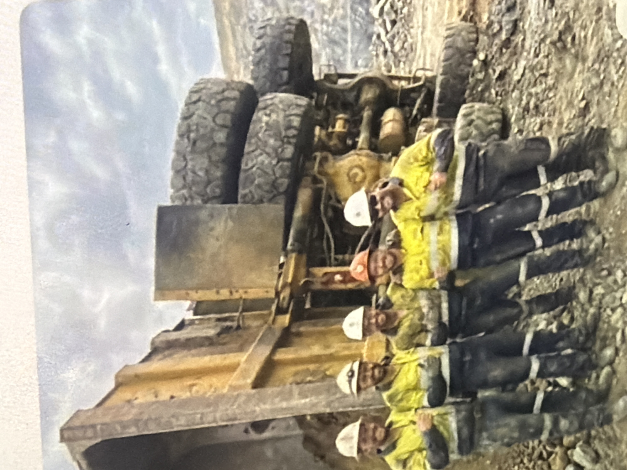 Six men in mining gear, including hard hats and reflective vests, pose for a photograph in front of a large, overturned yellow dump truck. The truck is on its side, with its massive tires visible. The men are standing on rough, rocky terrain, with a quarry or open-pit mine visible in the background. The sky is overcast.