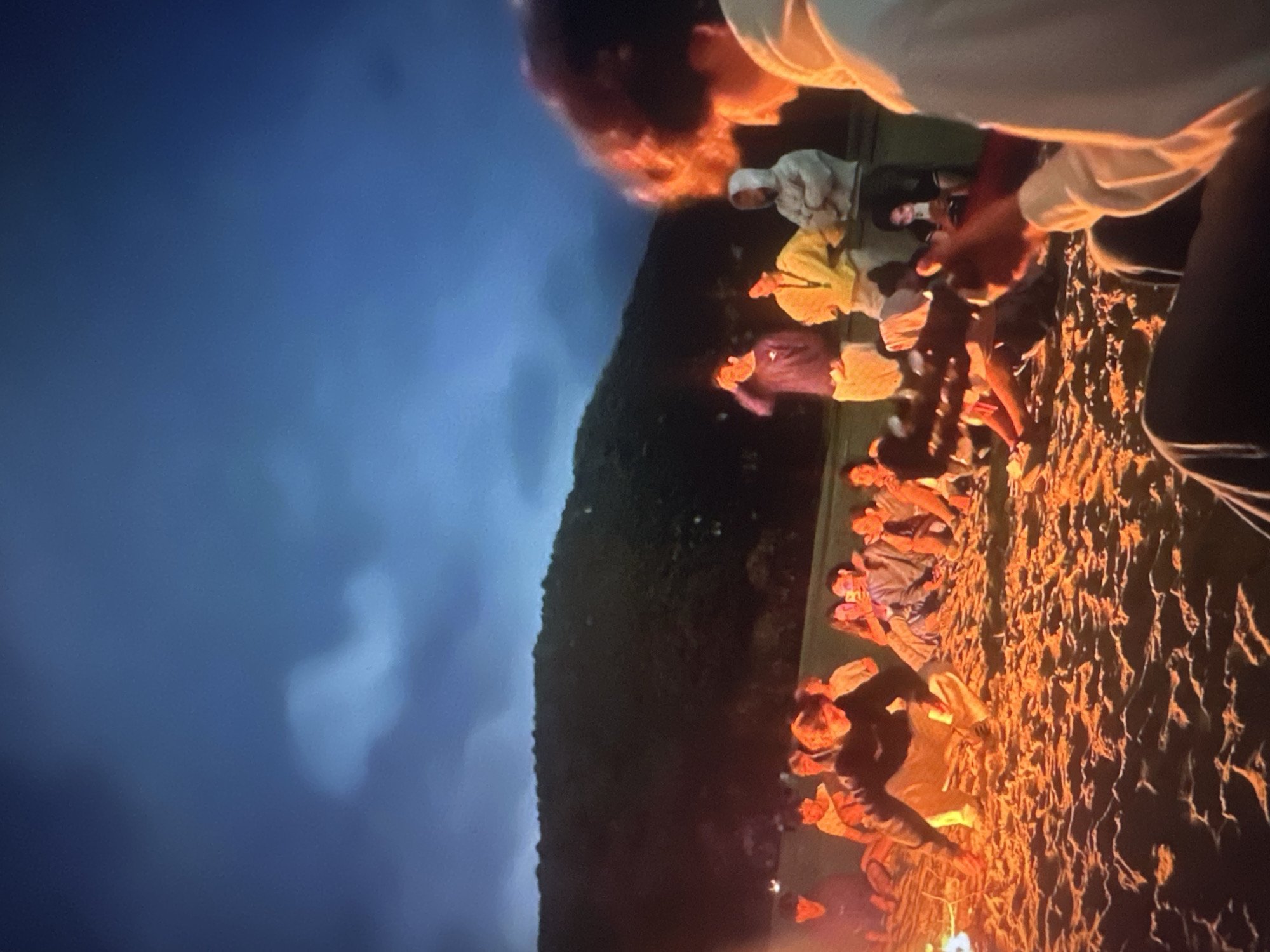 The image depicts a group of people gathered on a sandy beach at dusk, with a bonfire providing illumination. The sky is a deep blue with some clouds. In the foreground, a person with curly hair is holding a guitar, facing away from the camera. Behind them, several individuals are seated or standing around the bonfire, some appearing to be socializing. The beach slopes upwards towards a dark, tree-covered hillside in the background. The overall atmosphere suggests a casual outdoor gathering. There are no identifiable landmarks or elements that would specifically place this scene in Tallinn, Estonia.