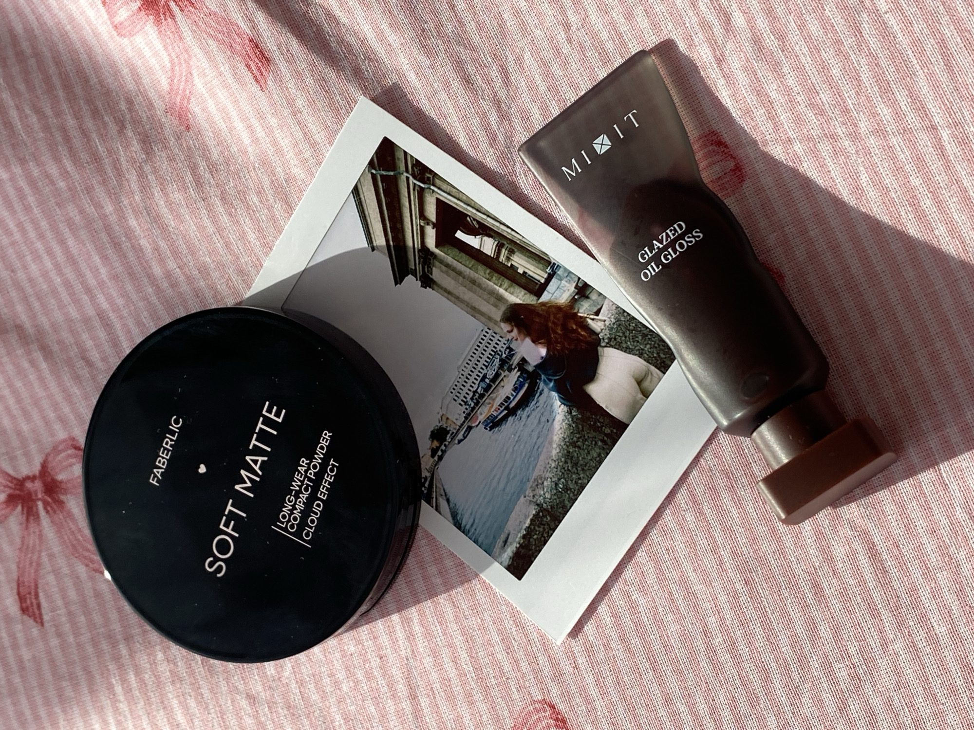 The image presents a still life composition featuring cosmetics and a photograph on a light pink fabric with a subtle pattern. A round, black compact powder case labeled "FABERLIC SOFT MATTE" is positioned on the left. To its right lies a square Polaroid photograph showing an outdoor scene with a woman standing along the Embankment of Izhevsk Pond in Izhevsk, Russia. Buildings are visible in the background, and the woman is standing near the pond edge. A dark tube of "MIXIIT GLAZED OIL GLOSS" is placed on the upper right, with its dispenser end pointed towards the viewer. The items are arranged casually on the fabric, suggesting a personal and informal setting.