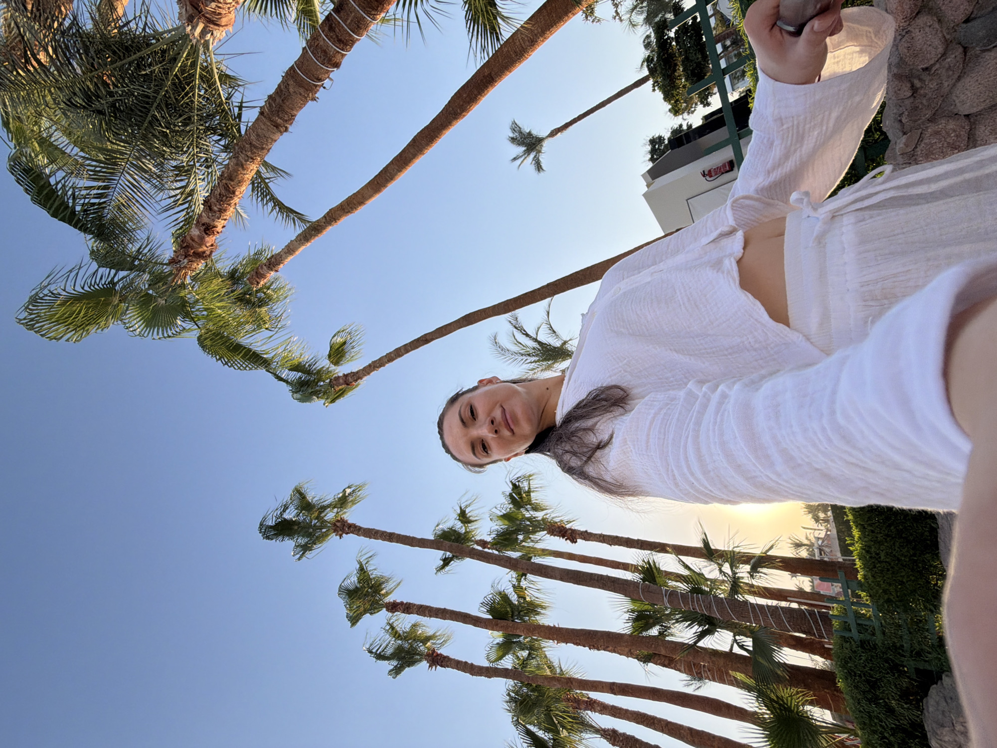 A woman stands in Sharm El Sheikh, Egypt, looking up at the camera. She is wearing a white outfit consisting of a cropped long-sleeved shirt and loose pants. The background features a clear blue sky and several tall palm trees. The sun is low in the sky, casting a warm glow. In the distance, there is a building and some greenery. The shot is taken from a low angle.