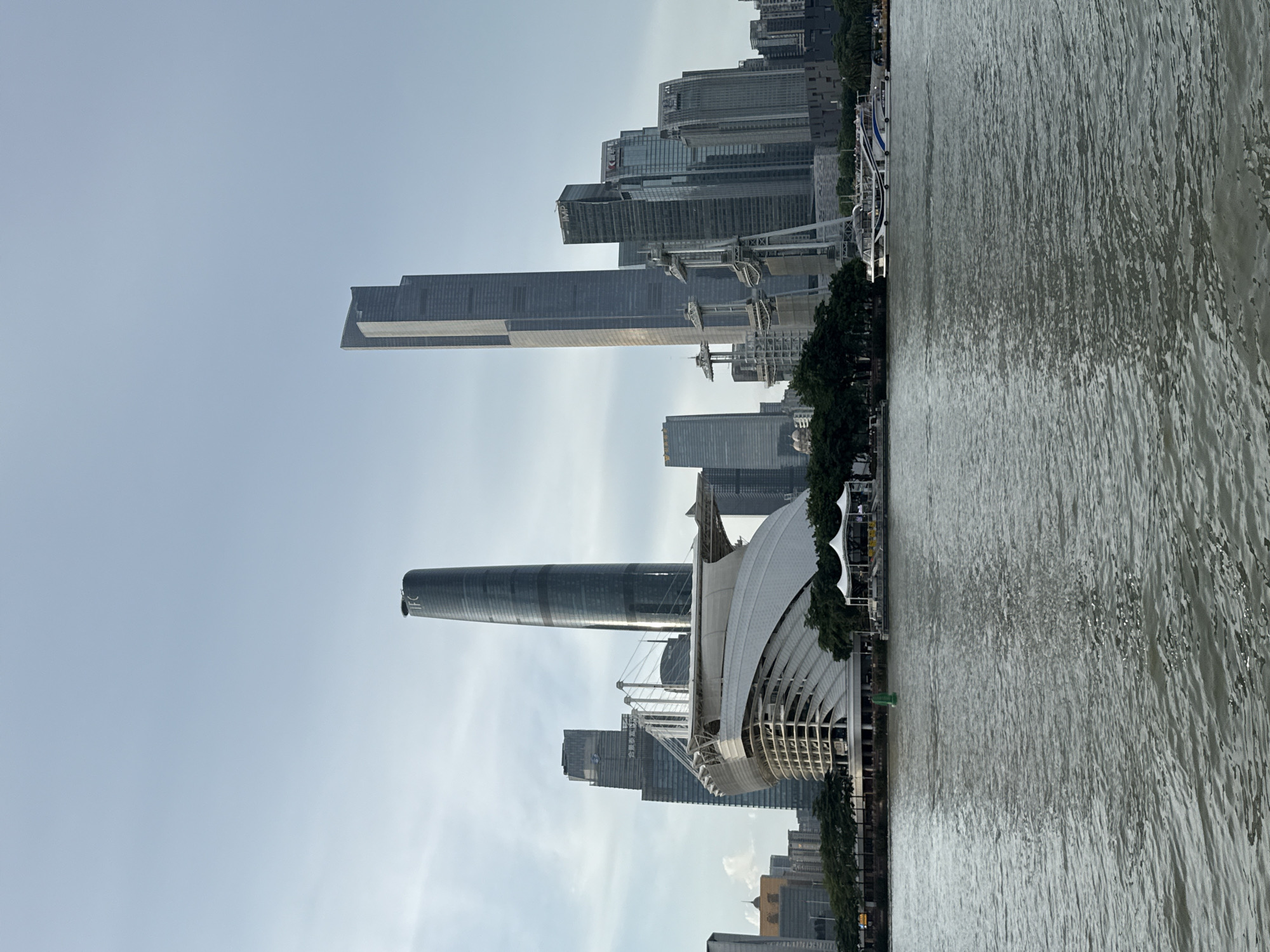 This is a photograph of the Guangzhou skyline, taken from across the Pearl River in Haizhu District, China. The image captures a selection of modern skyscrapers, including the iconic Canton Tower, a tall, slender building with a distinct curved design. In the foreground, the murky waters of the Pearl River are visible, with a white and blue boat sailing in the distance. The sky is overcast, with a soft, diffused light illuminating the scene. The composition is dominated by the imposing architecture of the city, showcasing the urban development and ambition of Guangzhou.