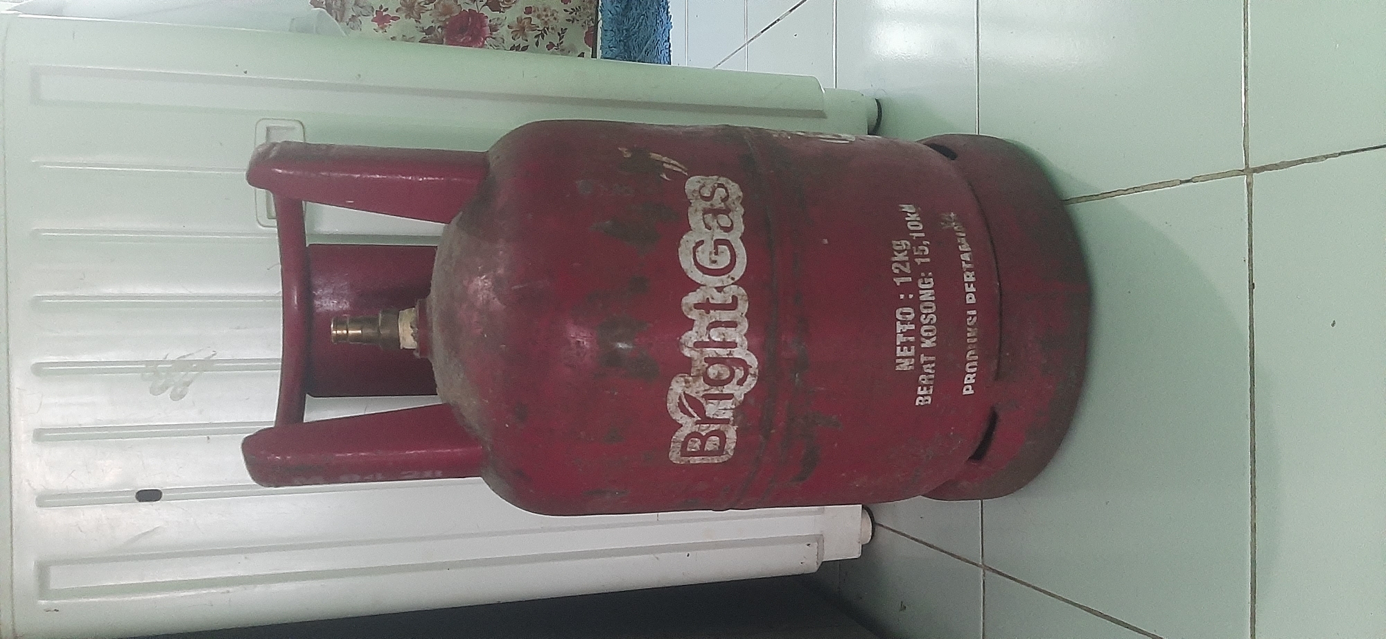 A red gas cylinder, identified as "Bright Gas," stands on a tiled floor in front of a white appliance. The cylinder has visible wear and tear, with rust spots and faded paint. Text on the cylinder indicates "NETTO: 12kg" and "BERAT KOSONG: 15,10kg." Below this, partially obscured, is text that appears to read "PRODUKSI PERTAMINA," suggesting it is a product of Pertamina, Indonesia's state-owned oil and natural gas corporation. The background appliance is white with vertical ribbed detailing. To the right of the cylinder, a floral curtain is visible, and a blue mat is on the floor. The lighting suggests an indoor setting. The image is a close-up shot, focusing on the gas cylinder. The presence of Pertamina branding strongly suggests this image was taken in Indonesia, and therefore potentially in the city of Jambi, Indonesia, as requested.