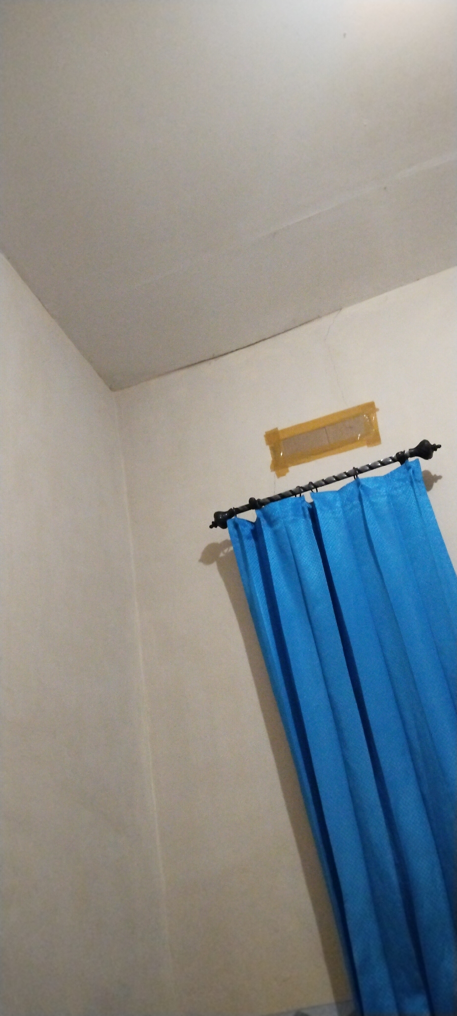 The image shows a corner of a room with a pale yellow wall and ceiling. A dark metal curtain rod extends horizontally across the wall, holding up a bright blue curtain that is partially drawn. Above the curtain rod, there is a rectangular vent covered with yellow tape. The lighting in the room is dim, casting shadows on the wall. The scene suggests an interior space, possibly a bedroom or living area, with the overall impression being one of simplicity. There are no people visible in the image. Without additional information or context, it is not possible to determine the specific location within the city of Duman, Indonesia.