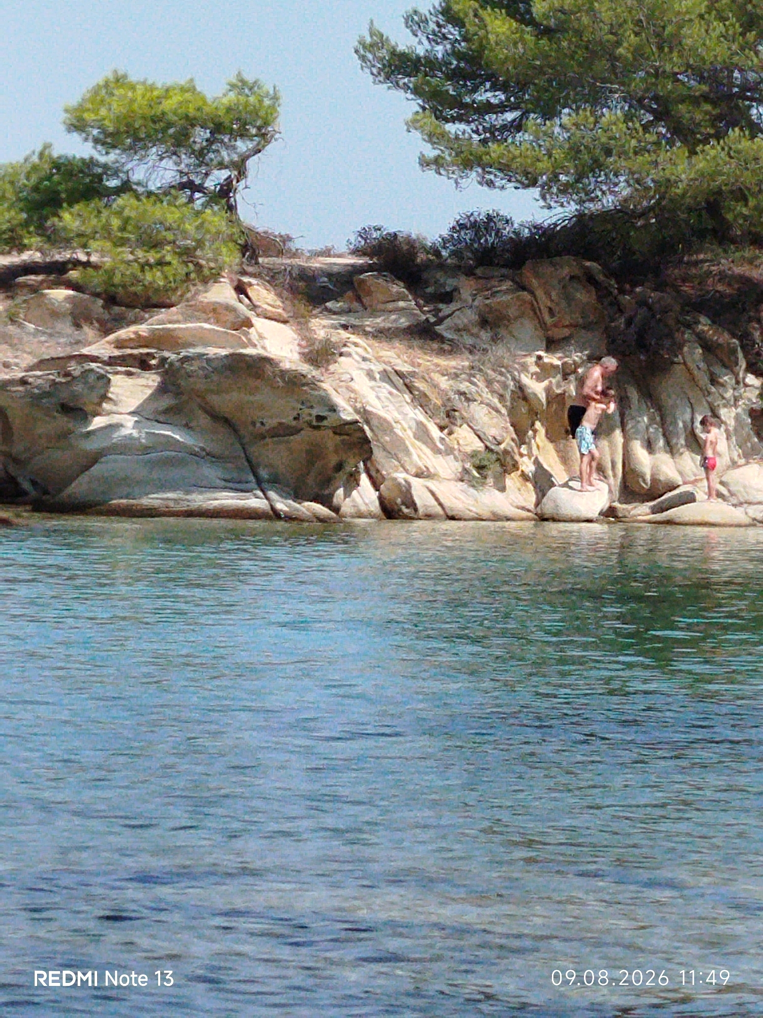 The image shows a rocky coastline with trees overlooking a body of water. Three people are visible near the shore. On the left, a man in dark shorts is holding onto a young boy in blue patterned shorts who is standing on a rock. To the right, a child in red shorts stands on another rock. The water in the foreground appears calm and clear. The rocky landscape suggests a natural environment, possibly a beach or a secluded cove. The date stamp at the bottom right indicates August 9, 2026, at 11:49 AM. The presence of the "REDMI Note 13" text at the bottom left suggests this is a photograph taken with a mobile device. The scene depicts a moment of leisure or recreation by the sea. The location is likely a coastal area in Sithonia Municipal Unit, Greece, known for its scenic beaches and rocky shores.