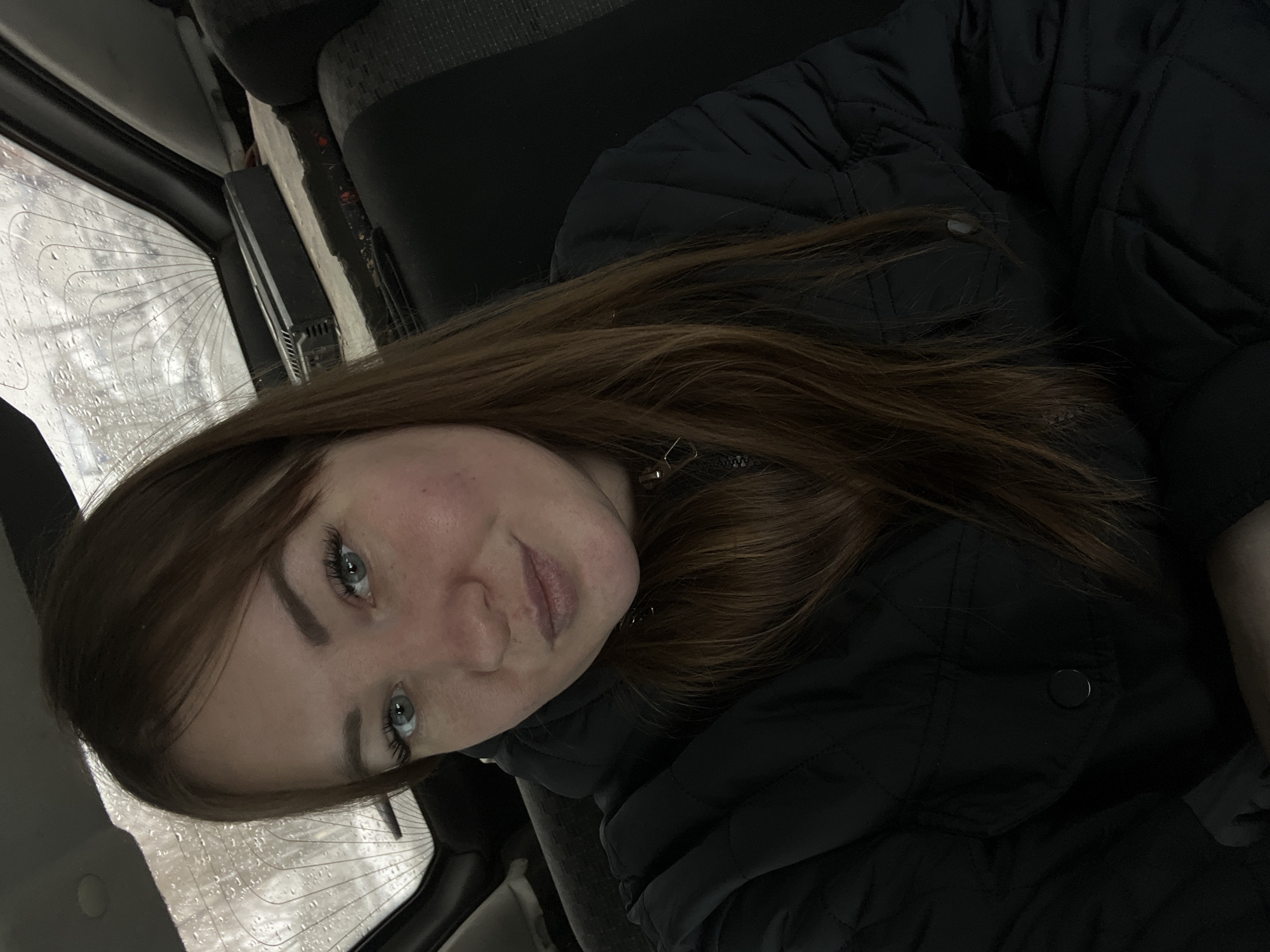 A young woman with light skin, light-colored eyes, and auburn hair is seated in what appears to be a car. She is wearing a black quilted jacket. Raindrops are visible on the car's window. The location is not discernible as Chaikovsky, Russia.