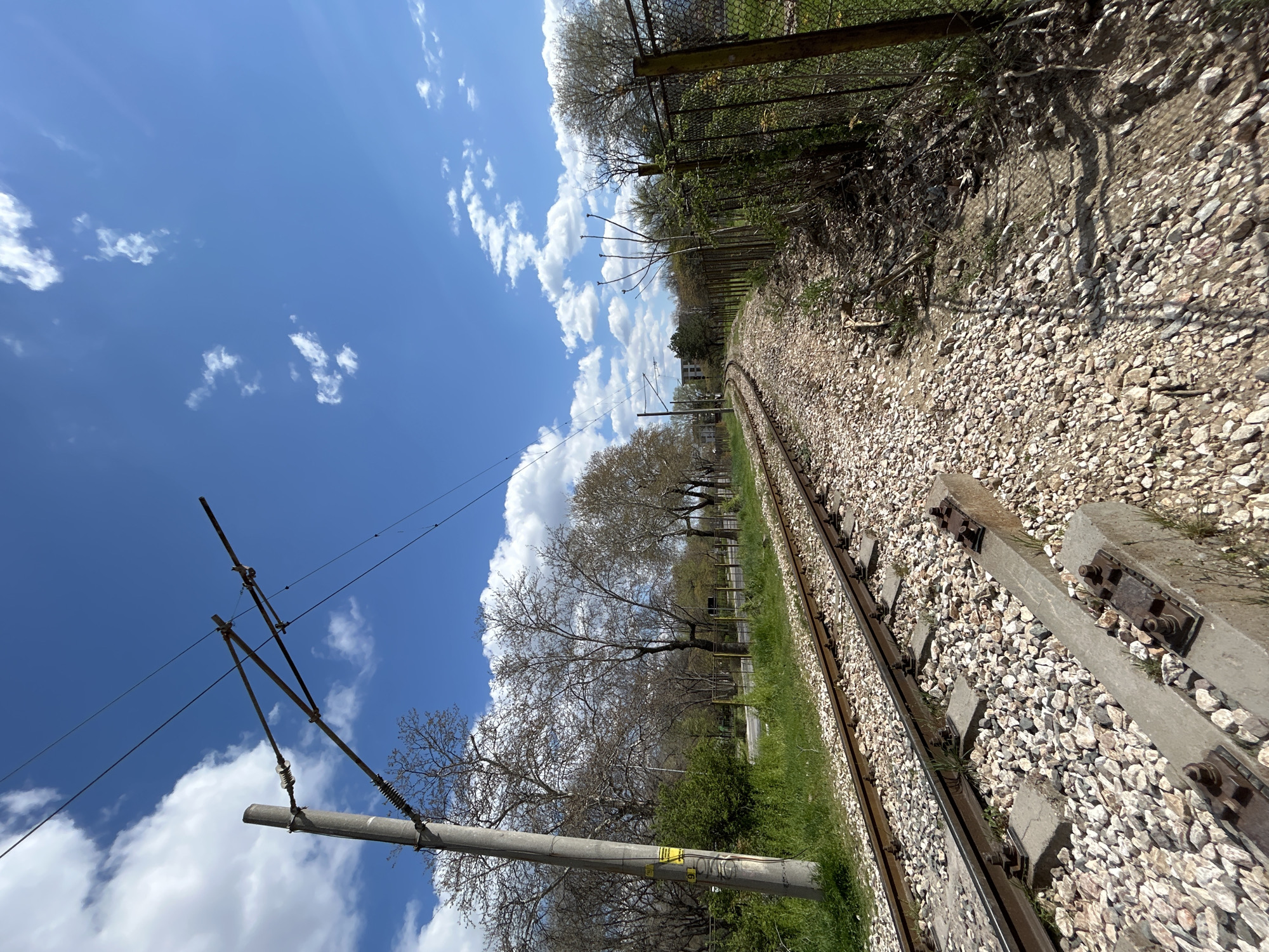 The image depicts a railway track extending into the distance under a blue sky with scattered clouds. The tracks are flanked by gravel ballast and concrete sleepers. Overhead electrical lines are visible, supported by concrete poles. To the right of the track is a grassy area, followed by a chain-link fence. Bare trees are visible along the edge of the tracks. The angle and perspective of the image suggest it was taken from a low vantage point, potentially from the track level itself. The location is identifiable as Plovdiv, Bulgaria, due to the specific infrastructure and typical landscape features.