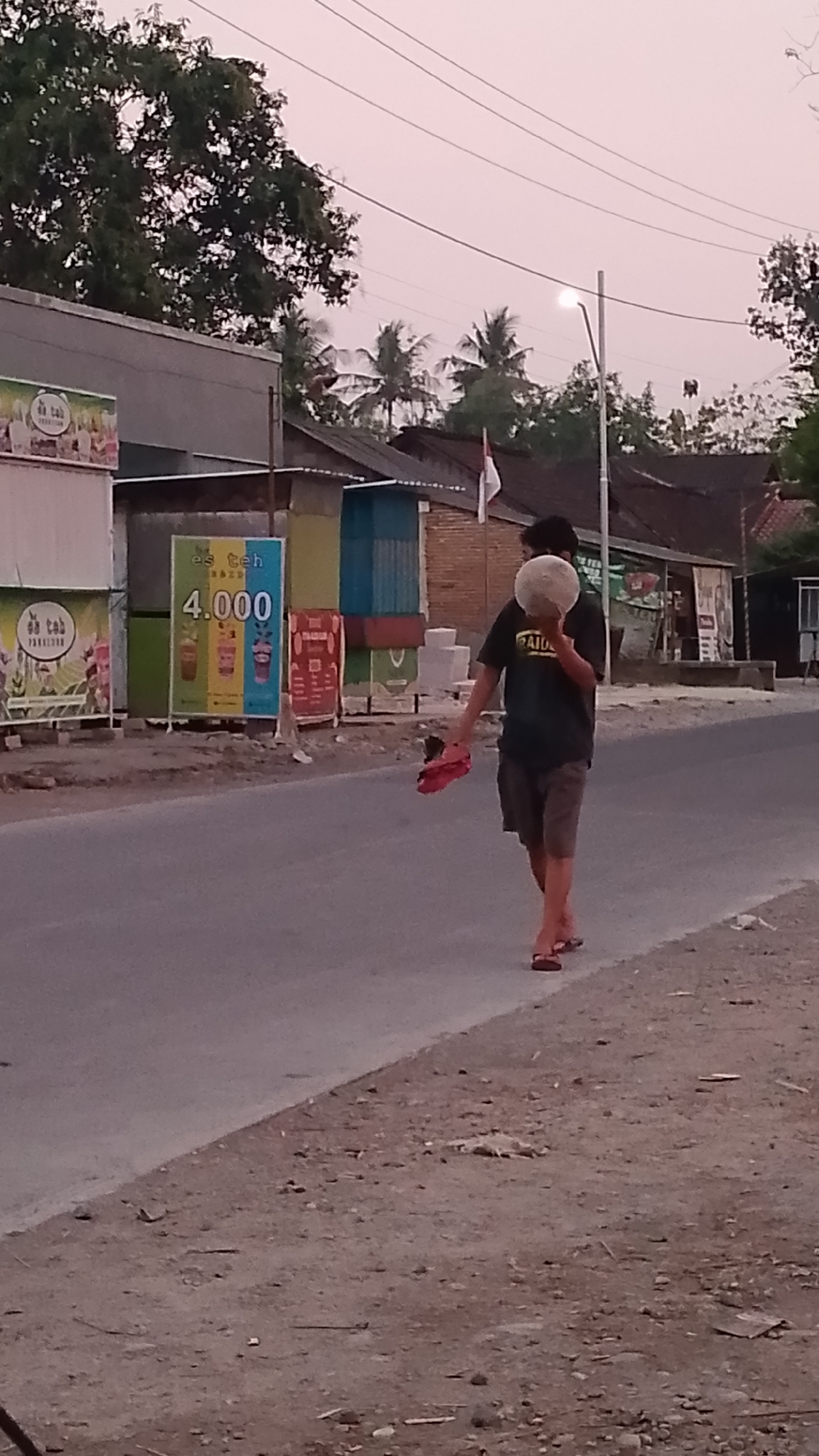 A man walks on the side of a road in Karanglangu, Indonesia, carrying a round, light-colored object, possibly a coconut, on his head. He is wearing a dark t-shirt and shorts, and is barefoot except for a pair of red flip-flops he is carrying in his right hand. The man is walking away from the camera, towards the right side of the frame. In the background, there are several small buildings with corrugated metal roofs and colorful advertisements on display. Palm trees and other foliage are visible, and the sky is a pale pink, suggesting it is either dawn or dusk. Power lines crisscross above the scene. The road appears to be asphalt, with a dirt and gravel shoulder on the right.